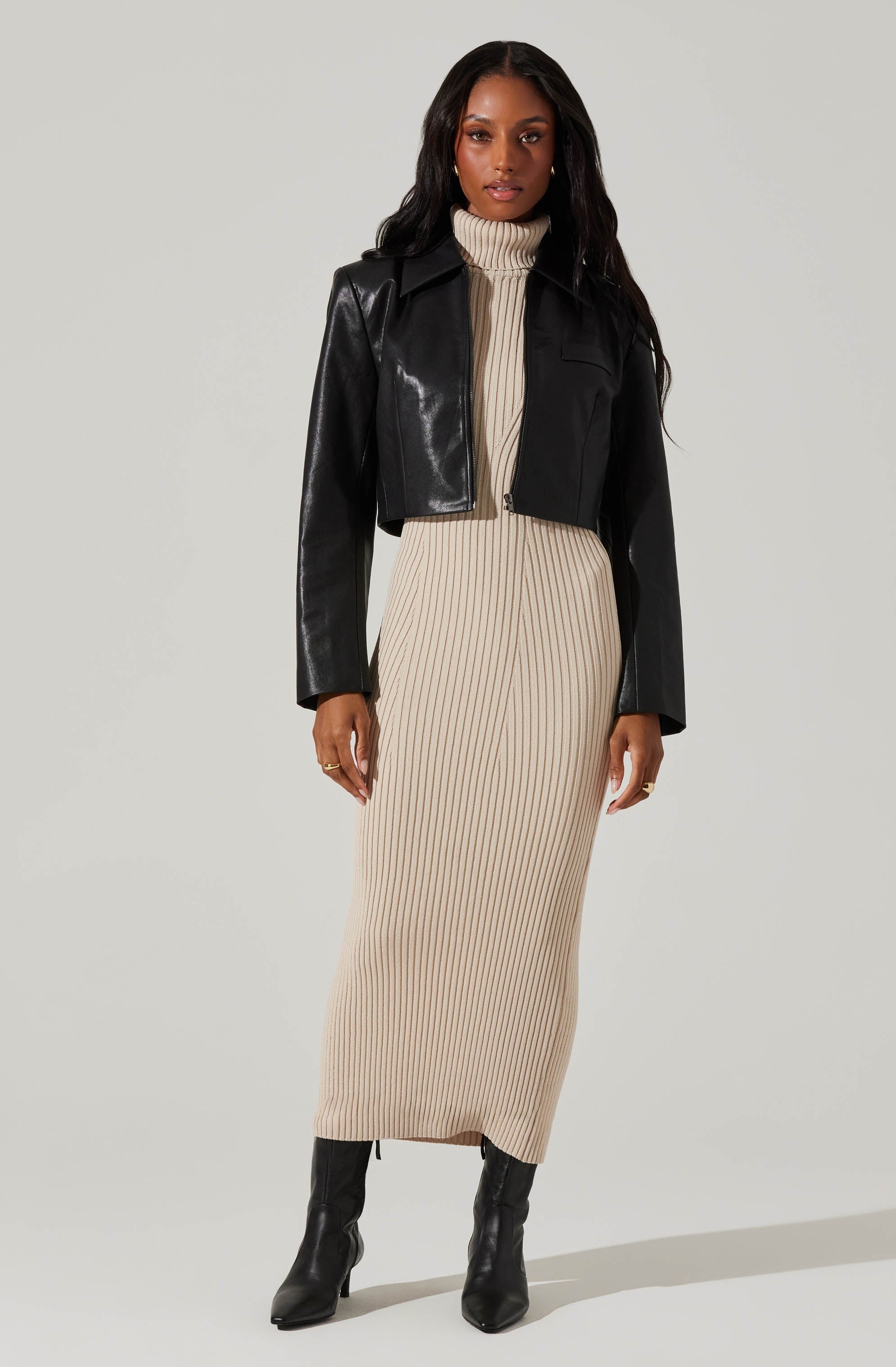 A woman is wearing a beige, ribbed, long-sleeve dress with a high neckline, paired with a black, cropped leather jacket and black high-heeled boots.