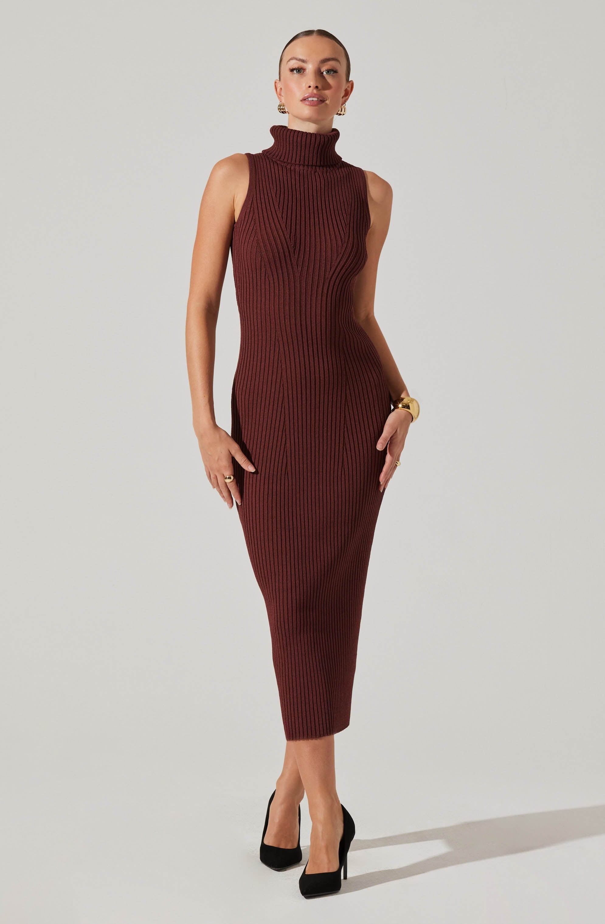 A woman is wearing a sleeveless, ribbed knit dress with a turtleneck in a deep burgundy color. The dress is form-fitting and extends to mid-calf length. She has paired the dress with black high-heeled shoes, gold earrings, and a gold bracelet.