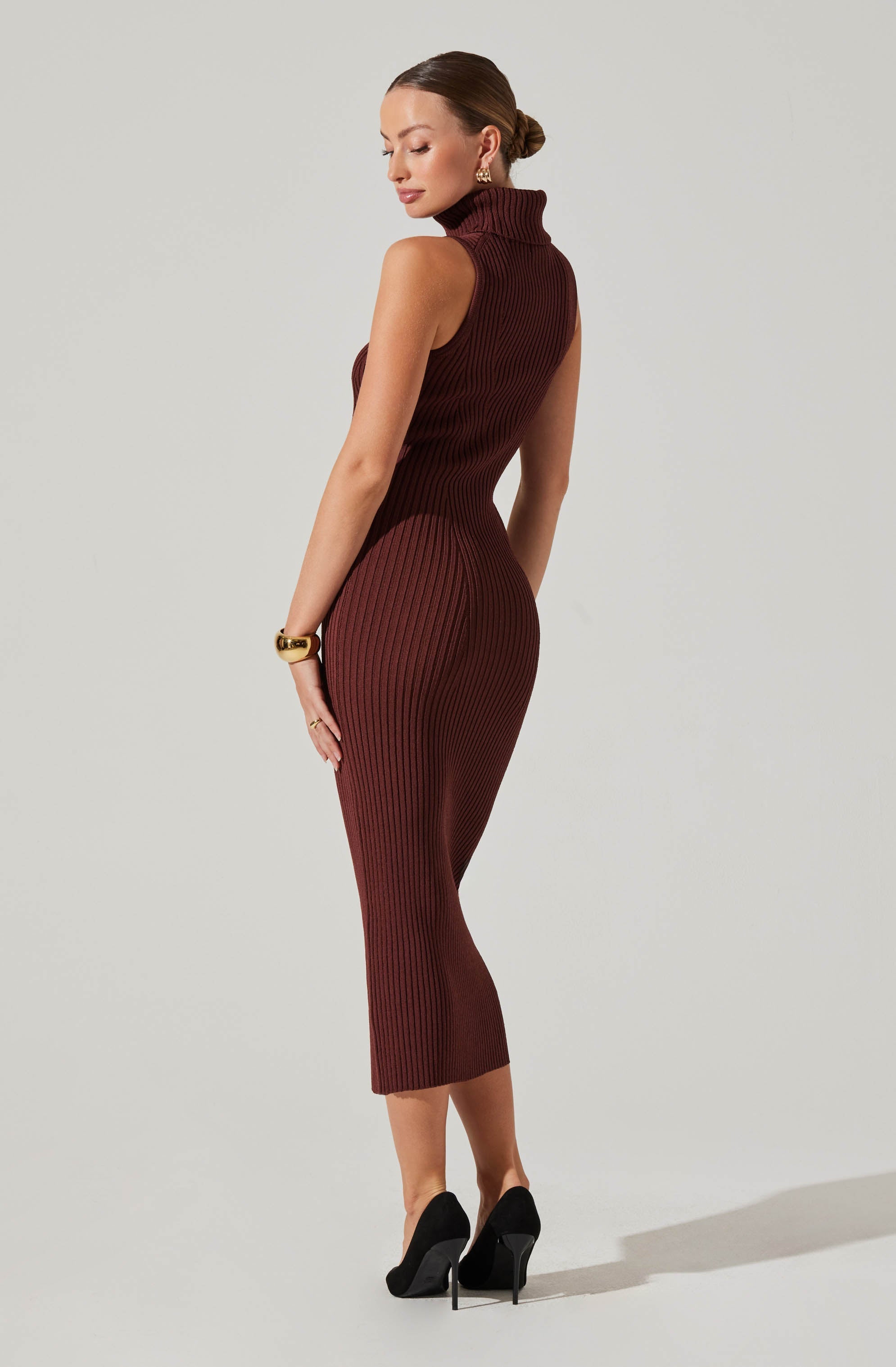 A woman is wearing a sleeveless, ribbed, turtleneck dress in a deep burgundy color that fits snugly and extends to mid-calf length. She accessorizes with gold earrings, a gold bracelet, and rings. She is also wearing black high-heeled shoes.