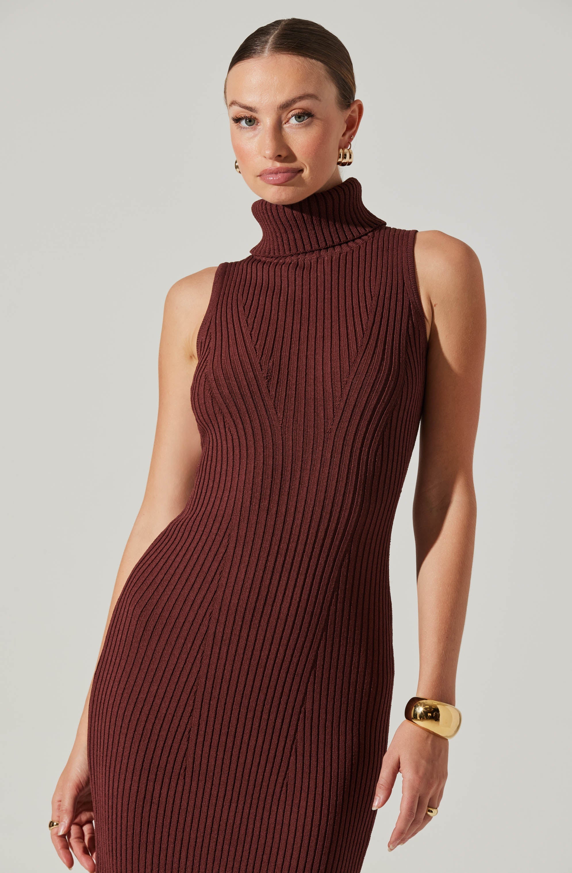 A woman is wearing a sleeveless, ribbed knit dress in a deep reddish-brown color, featuring a high turtleneck collar and vertical ribbing details running throughout the dress.