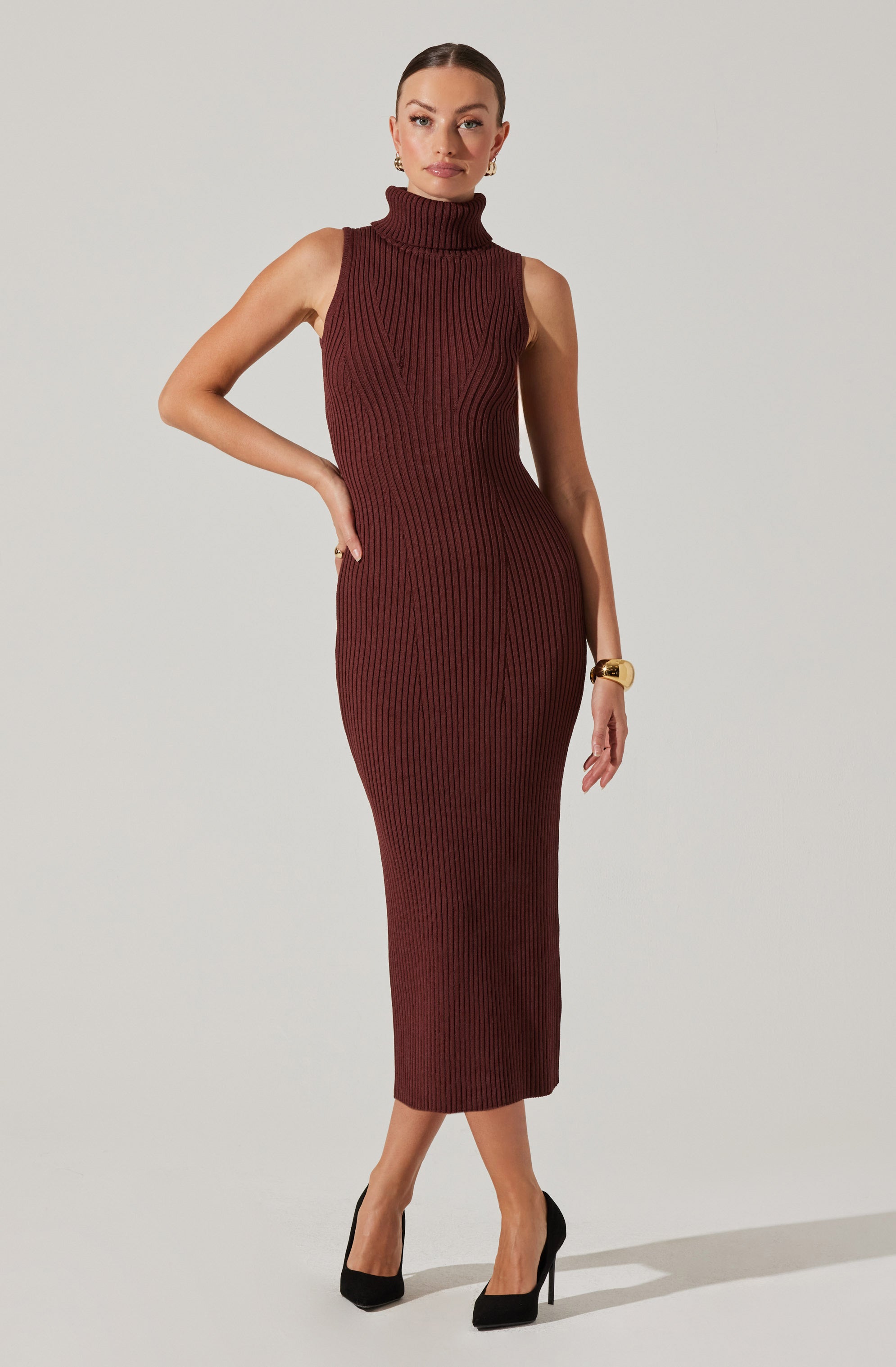 A woman is wearing a sleeveless, high-neck, ribbed knit dress that is fitted and extends to mid-calf length. The dress is a deep burgundy color with vertical ribbing. She has her right hand on her hip and is accessorizing with gold hoop earrings and a gold bracelet. She is wearing black high-heeled shoes.