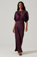 A woman is wearing a deep purple satin dress with a loose-fitting, draped top that has wide sleeves and a keyhole neckline, paired with a fitted, high-waisted, long skirt that reaches the floor. She is also wearing black open-toe heels.