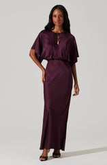 A woman is wearing a deep purple satin evening gown featuring a smooth, shiny fabric. The dress has a loose-fitting, draped bodice with a keyhole neckline and short, wide sleeves. The skirt is fitted at the waist and extends to the floor with a subtle side slit.
