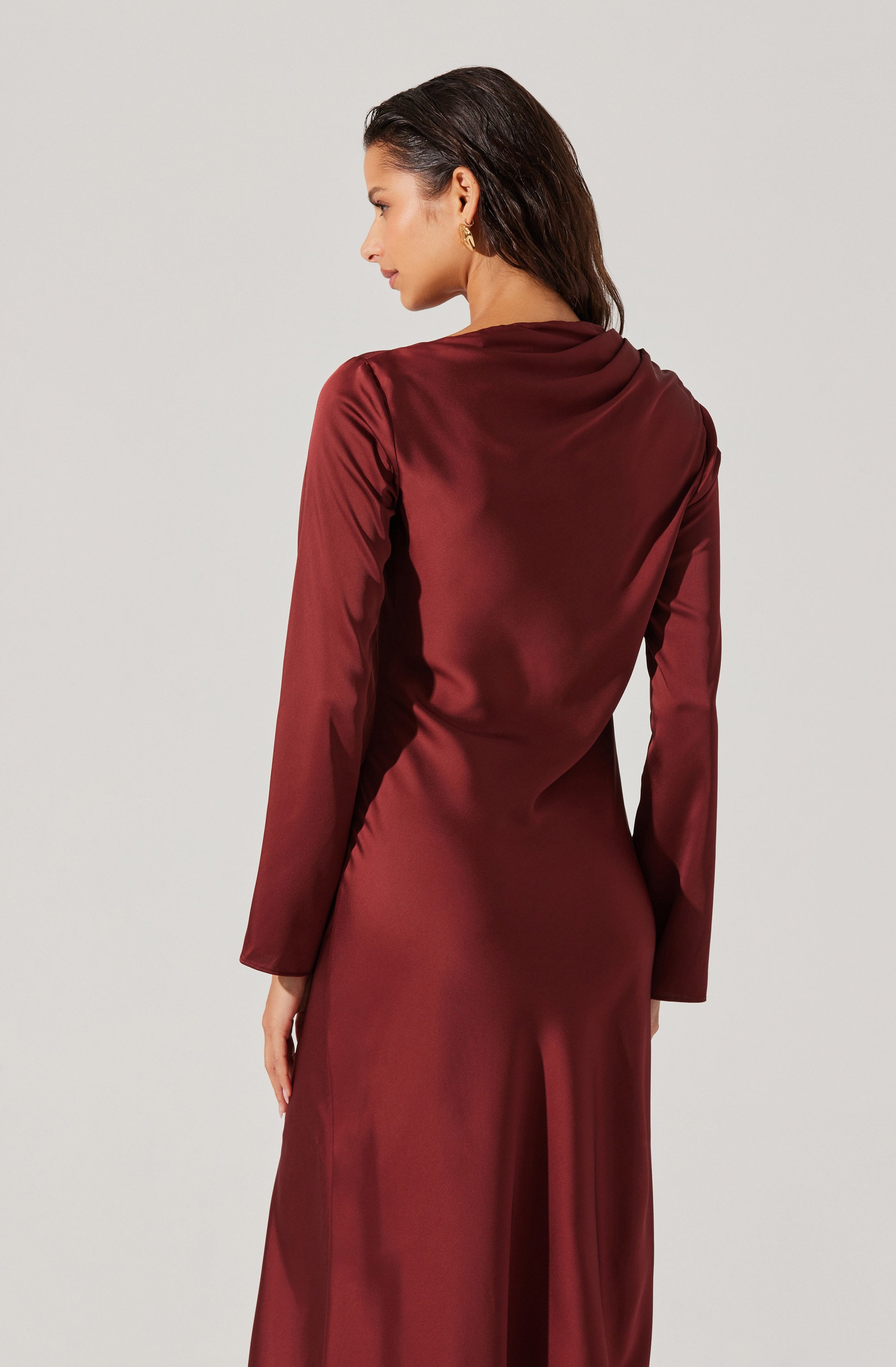 A woman is wearing a long, elegant, burgundy satin dress with long sleeves. The dress has a smooth, shiny finish and a fitted silhouette that drapes gently over her body. The fabric appears luxurious and soft, with a subtle sheen that catches the light.