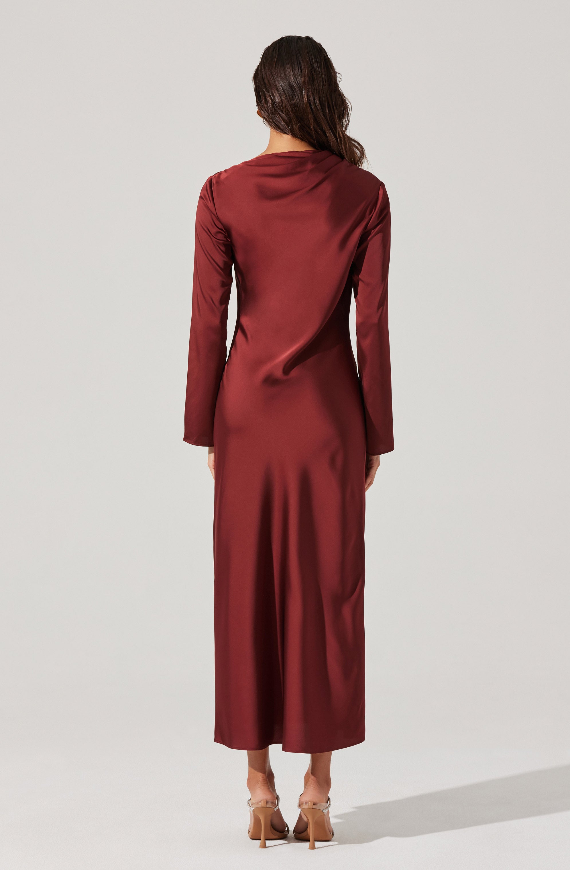 A woman is wearing a long, elegant, burgundy satin dress with long sleeves. The dress has a smooth, shiny texture and reaches down to her ankles. She is also wearing tan high-heeled shoes.
