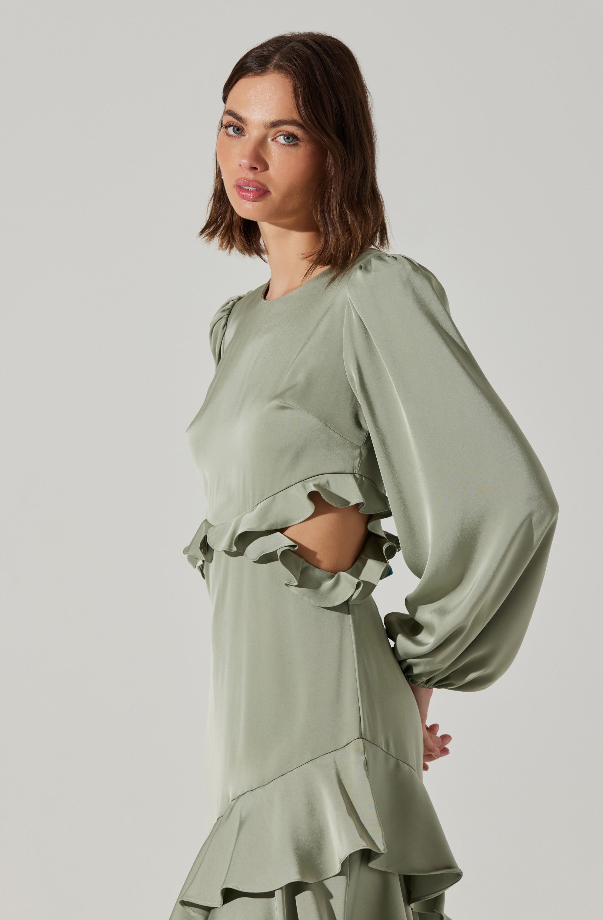 A woman is wearing a light green satin dress with long, puffed sleeves. The dress features ruffled details at the waist and hem, creating a layered, flowy appearance. The fabric appears smooth and glossy.