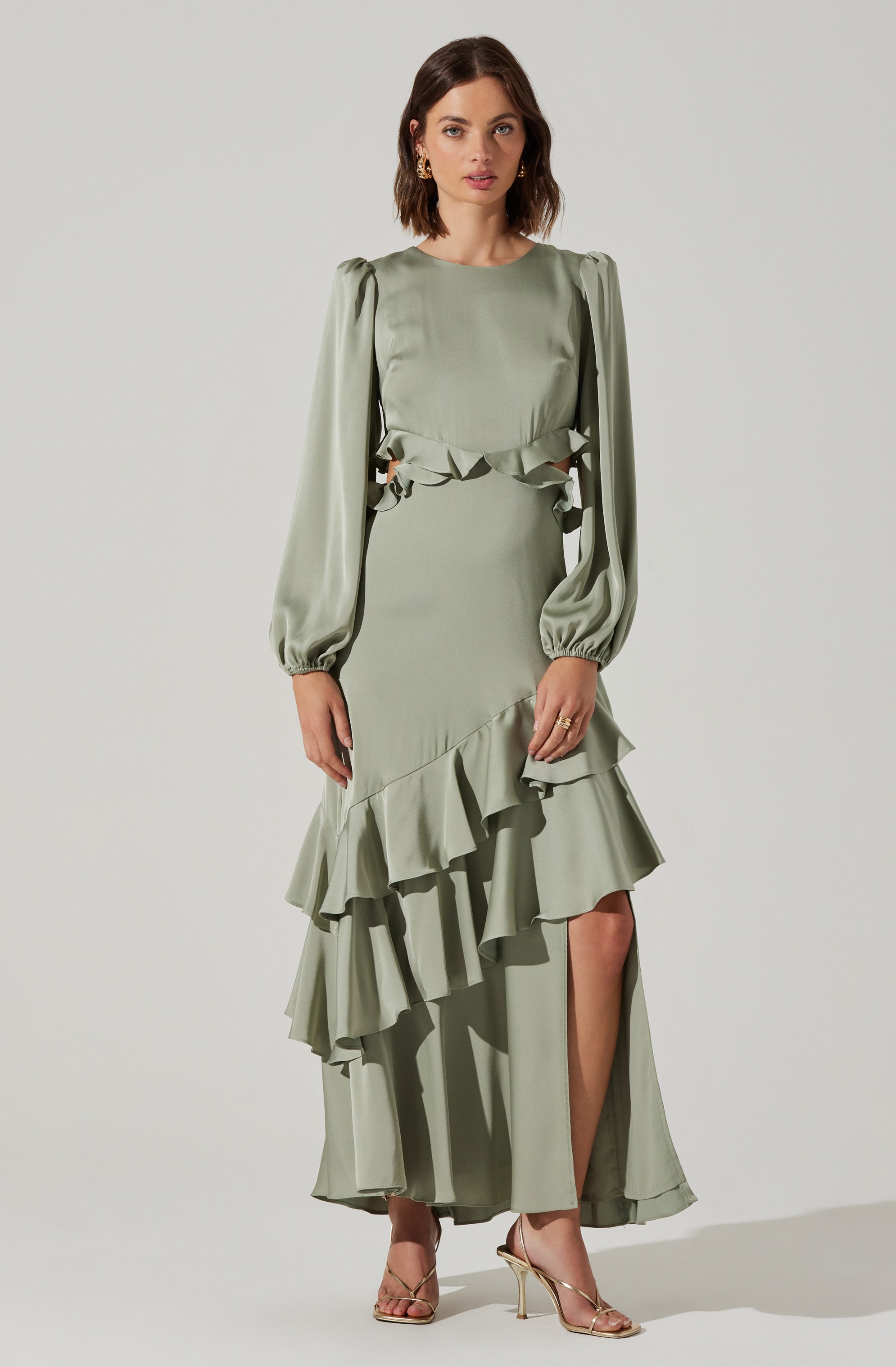 A woman is wearing a long, pale green dress with puffed long sleeves that are gathered at the wrists. The dress features cut-out details on the sides of the waist with ruffled accents, and a layered, ruffled hemline that cascades asymmetrically down the front and back, revealing a high slit on one side. She is also wearing gold high-heeled sandals and gold hoop earrings.