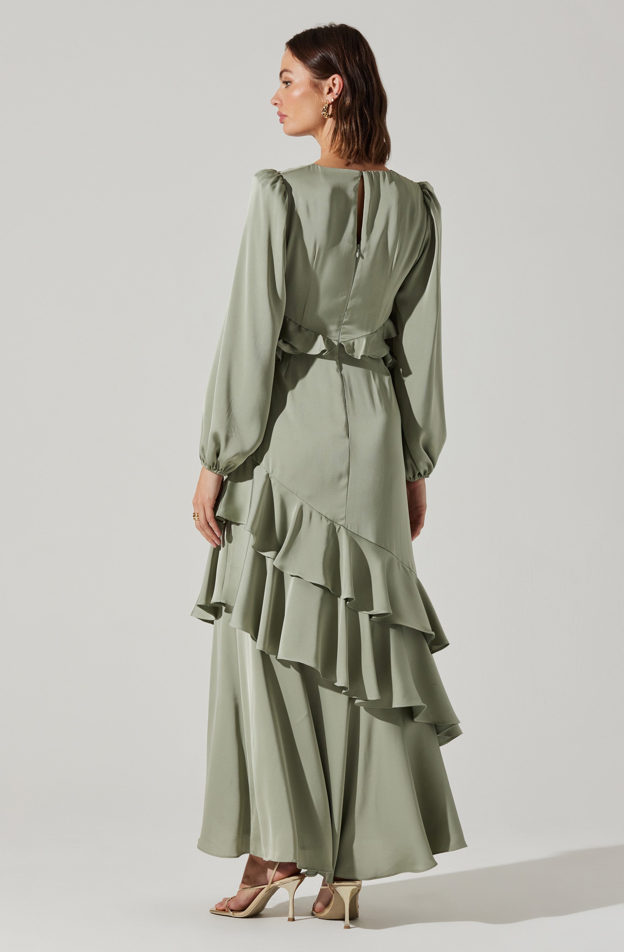 A woman is wearing a light green, long-sleeved dress with puffed sleeves, a keyhole back with a small button closure, a waist tie, and a layered, ruffled skirt that extends to the floor. She has on beige high-heeled shoes and gold hoop earrings.