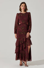 A woman is wearing a long, burgundy dress with an intricate floral pattern in a darker shade. The dress features puffed long sleeves with elastic cuffs, a ruffled front detail at the waist, and a high slit on one side revealing her leg. She is also wearing black strappy high-heeled sandals.