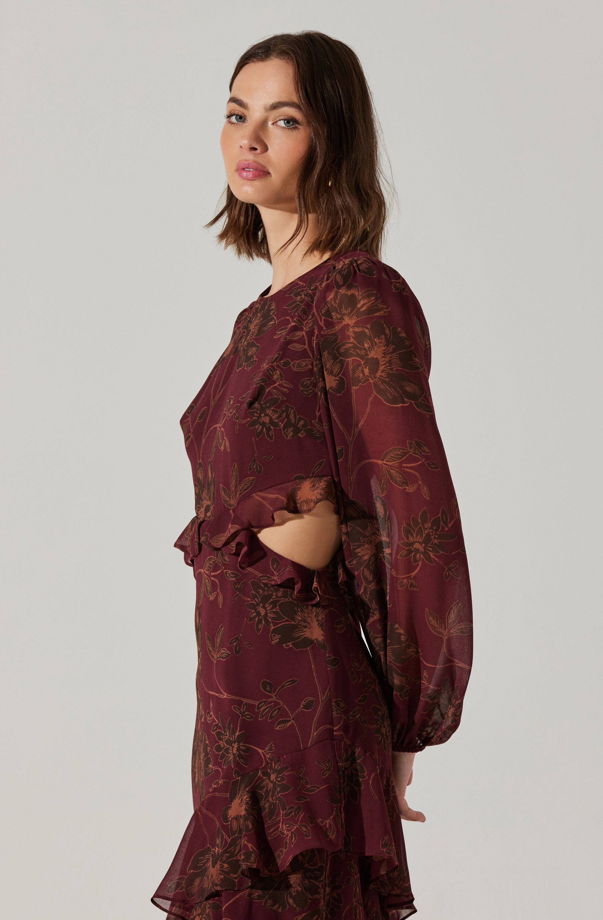 A woman is wearing a long-sleeved, burgundy dress with a floral pattern in shades of brown and orange. The dress features gathered fabric at the cuffs and a ruffled hem that adds a decorative touch.