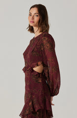 A woman is wearing a long-sleeved, burgundy dress with a floral pattern in shades of brown and orange. The dress features gathered fabric at the cuffs and a ruffled hem that adds a decorative touch.