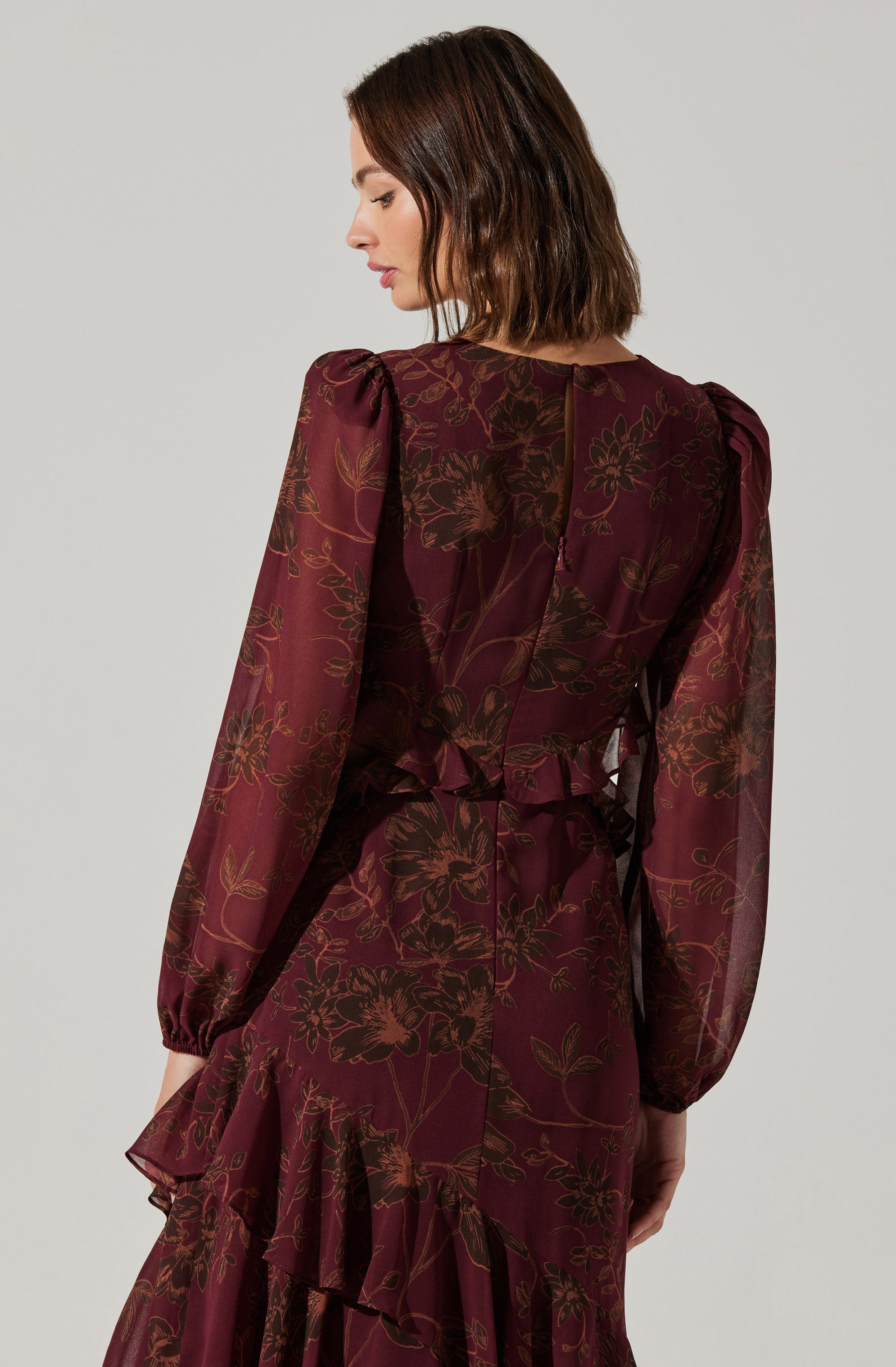A woman is wearing a burgundy dress with a floral pattern in shades of dark brown. The dress has long, sheer puffed sleeves, a fitted waist, and a ruffled hemline. The fabric appears lightweight and semi-transparent.