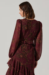 A woman is wearing a burgundy dress with a floral pattern in shades of dark brown. The dress has long, sheer puffed sleeves, a fitted waist, and a ruffled hemline. The fabric appears lightweight and semi-transparent.