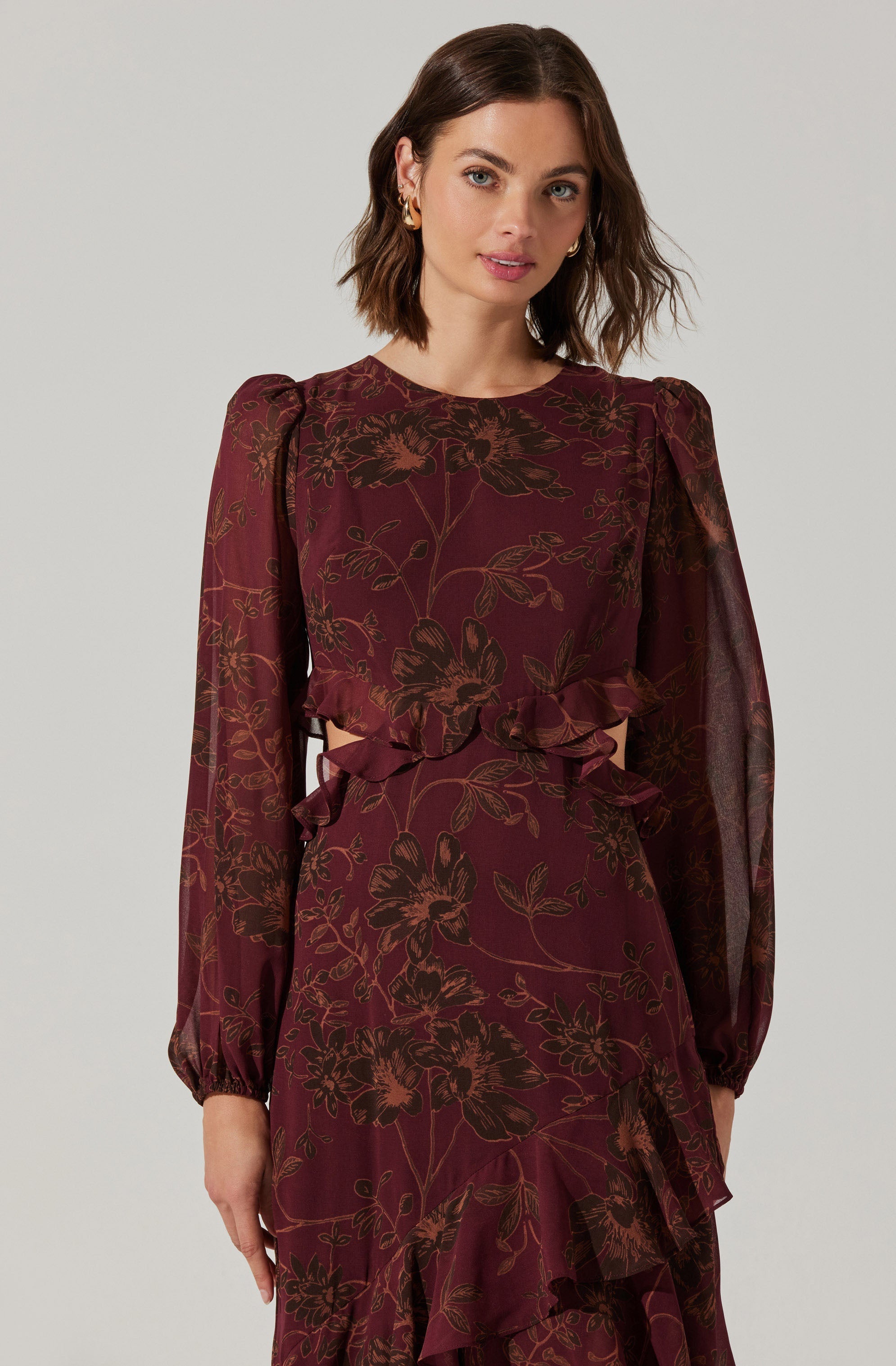 A woman is wearing a long-sleeved, burgundy dress with a floral pattern in shades of brown. The dress features puffed shoulders, sheer long sleeves, and ruffle details at the waist and the hem of the skirt.