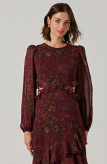 A woman is wearing a long-sleeved, burgundy dress with a floral pattern in shades of brown. The dress features puffed shoulders, sheer long sleeves, and ruffle details at the waist and the hem of the skirt.
