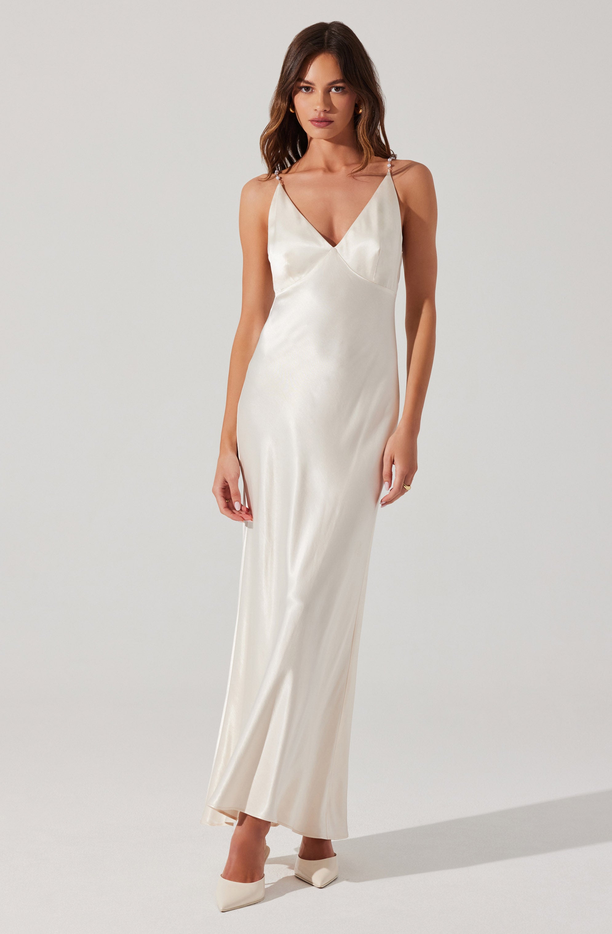 A woman is wearing a long, elegant satin dress in an ivory or champagne color. The dress features a V-neckline, thin spaghetti straps, and a fitted silhouette that flows down to the floor. She is also wearing matching pointed-toe heels.