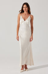 A woman is wearing a long, elegant, ivory satin gown with a deep V-neckline. The dress features thin, beaded spaghetti straps and a fitted silhouette that flows down to the floor. She is also wearing matching ivory shoes with a pointed toe.