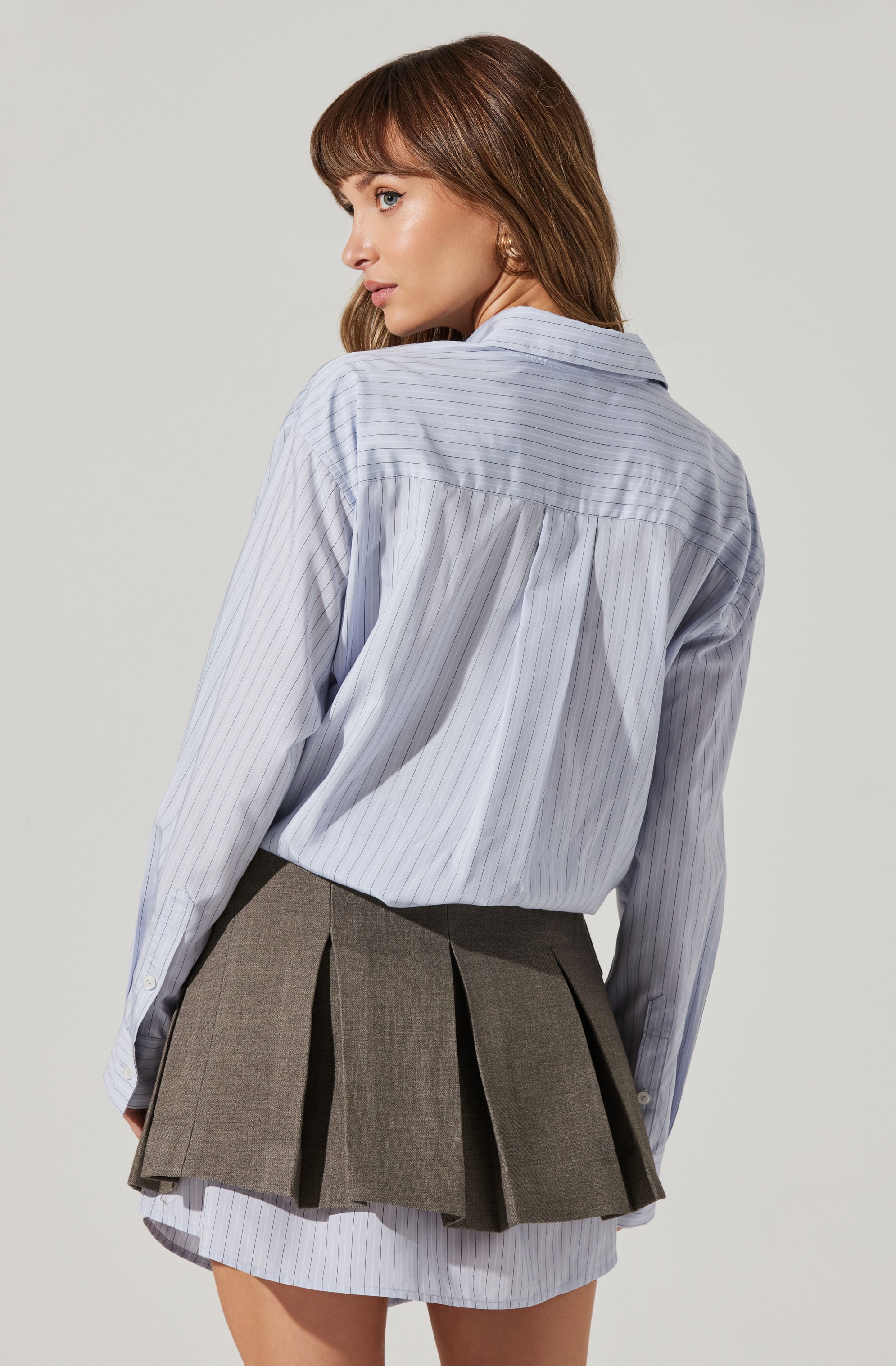 A woman is wearing a light blue, long-sleeved, collared shirt with thin vertical white stripes. The shirt is slightly oversized and tucked into a layered, pleated gray skirt. The skirt is short, with a portion of a matching striped fabric visible at the hem underneath the pleated layer.