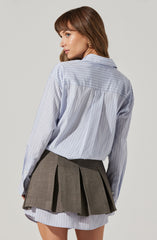 A woman is wearing a light blue, long-sleeved, collared shirt with thin vertical white stripes. The shirt is slightly oversized and tucked into a layered, pleated gray skirt. The skirt is short, with a portion of a matching striped fabric visible at the hem underneath the pleated layer.