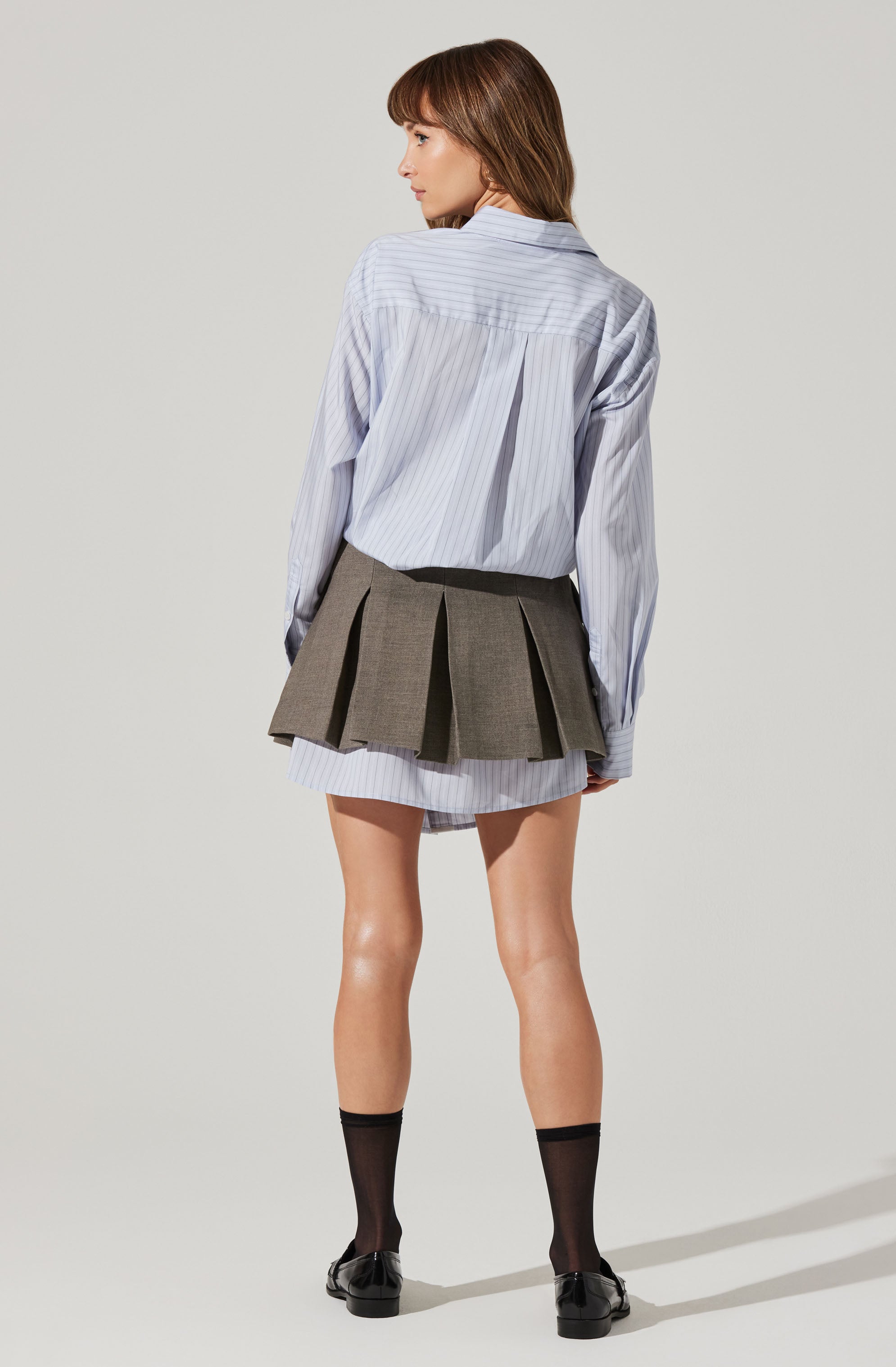 A woman is wearing an oversized light blue and white striped button-down shirt with long sleeves, paired with a gray pleated mini skirt layered over the shirt. She has black mid-calf socks and black loafers.