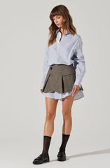 A woman is wearing a light blue button-up shirt with thin white vertical stripes, tucked into a high-waisted gray pleated skirt with black buttons and large front pockets. She has on black socks and black loafers with a small heel.