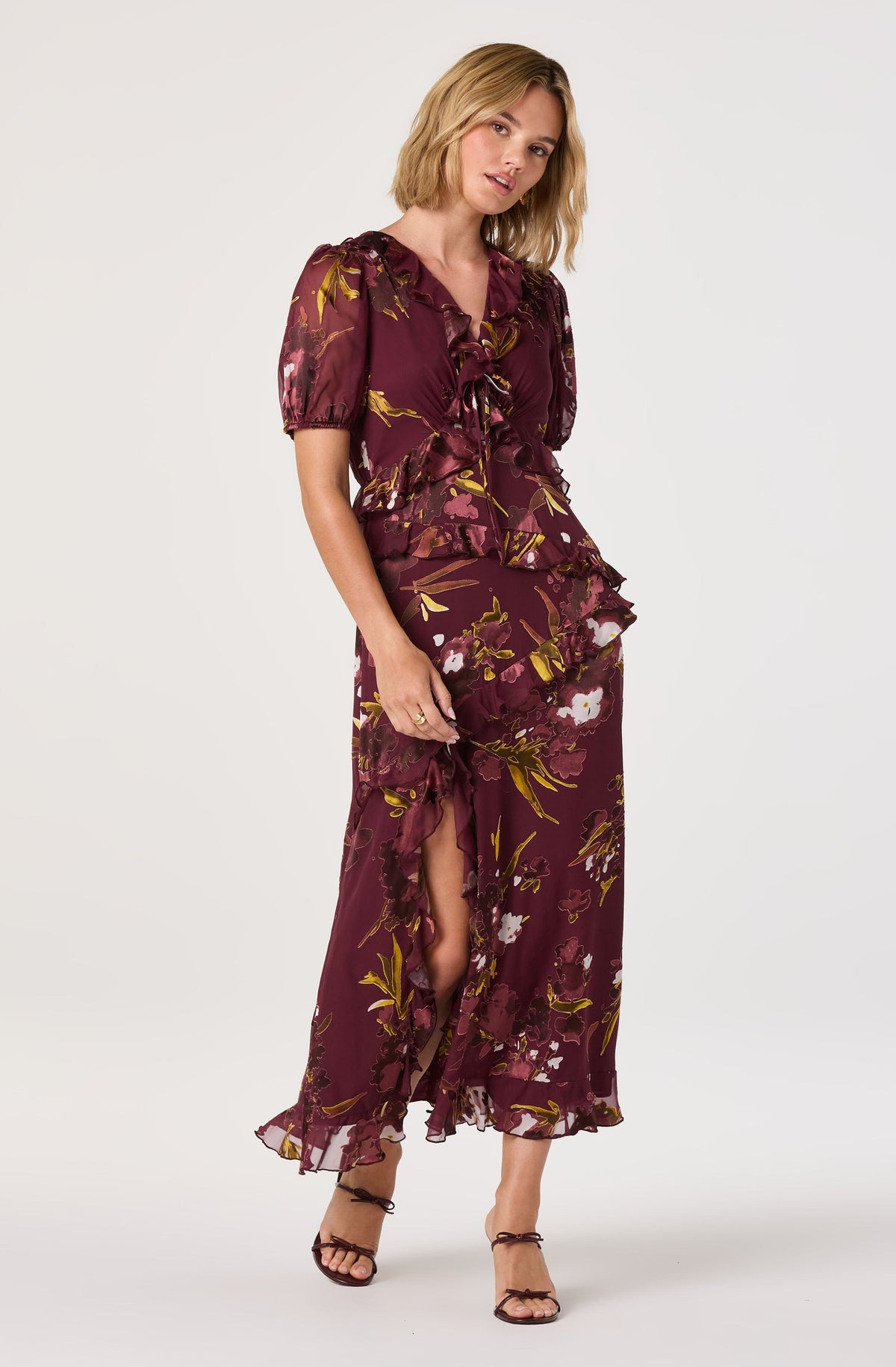 A woman is wearing a matching two-piece outfit with a deep burgundy background and a floral pattern featuring white, pink, and yellow flowers with green leaves. The top has short puffed sleeves, a V-neckline with ruffles, and a tie detail at the front. The skirt is long and flowing with a slit up one side, revealing part of her leg. The fabric appears lightweight and sheer. She is also wearing strappy black high-heeled sandals.