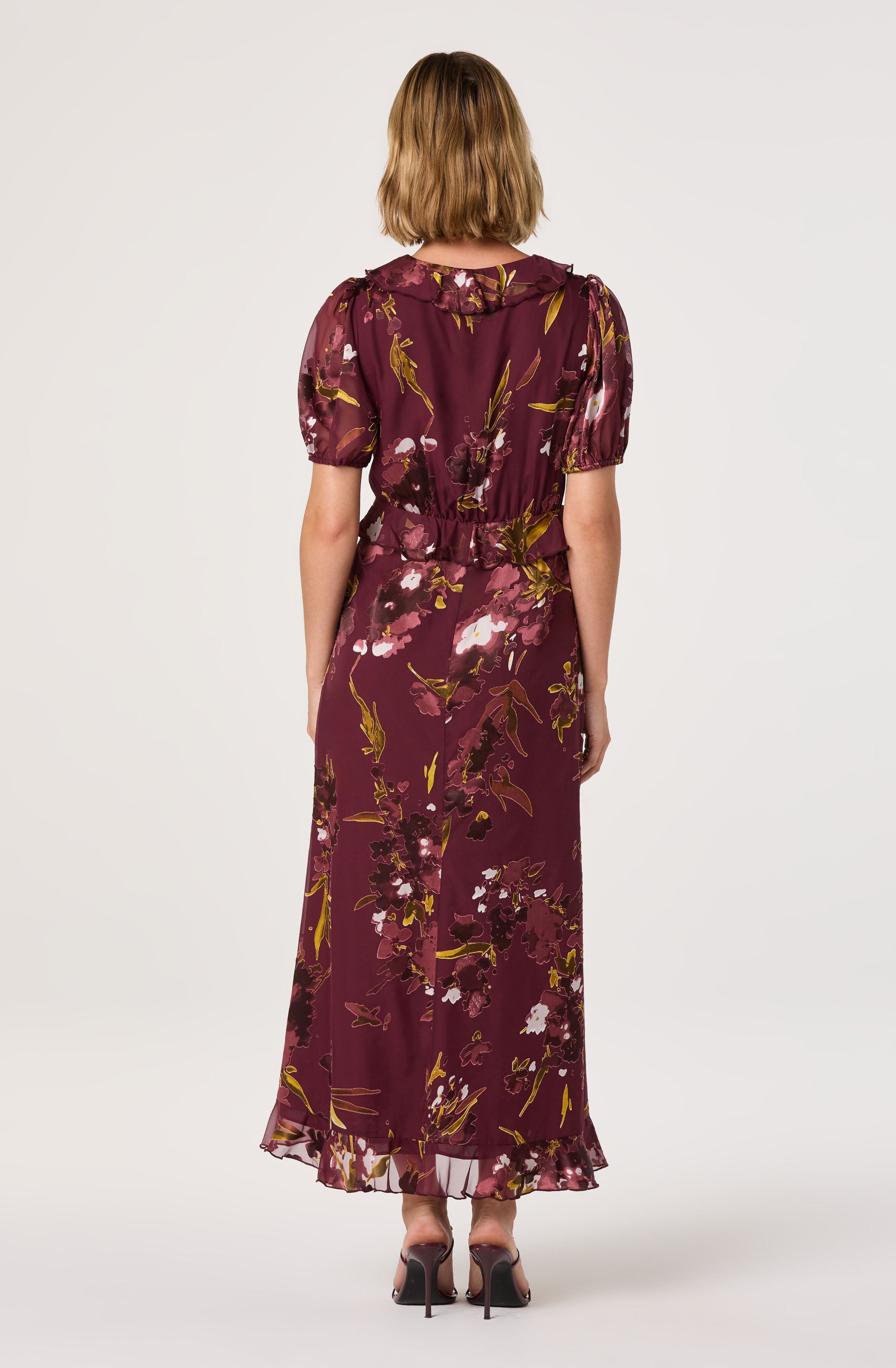 A woman is wearing a long, flowing dress with a floral pattern in shades of maroon, pink, green, and white. The dress has short, puffed sleeves, a fitted waist, and a ruffled hem at the bottom.