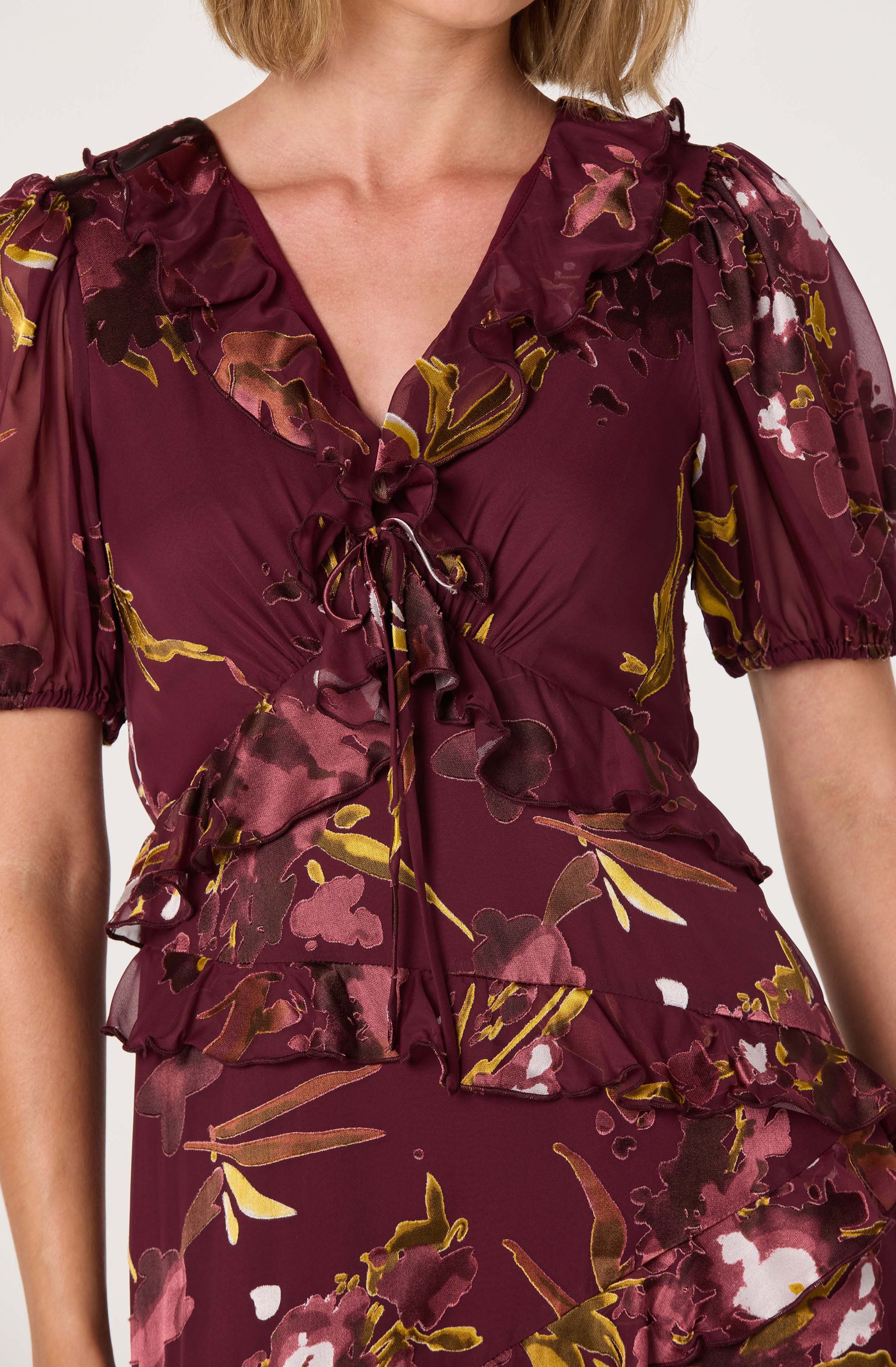 A woman is wearing a burgundy dress with a floral pattern in shades of pink, white, and gold. The dress features puffed short sleeves, a ruffled V-neckline, and ruffle details across the bust and waist. It is made of a shiny, silky fabric that drapes smoothly and has a tied bow at the center of the neckline.