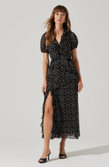 A woman is wearing a black dress with a white polka dot pattern. The dress features a V-neckline, puffed short sleeves, and a tiered ruffle detail that runs down the front and around the hem. It has a high slit on one side, revealing her leg. She is also wearing black high-heeled sandals.