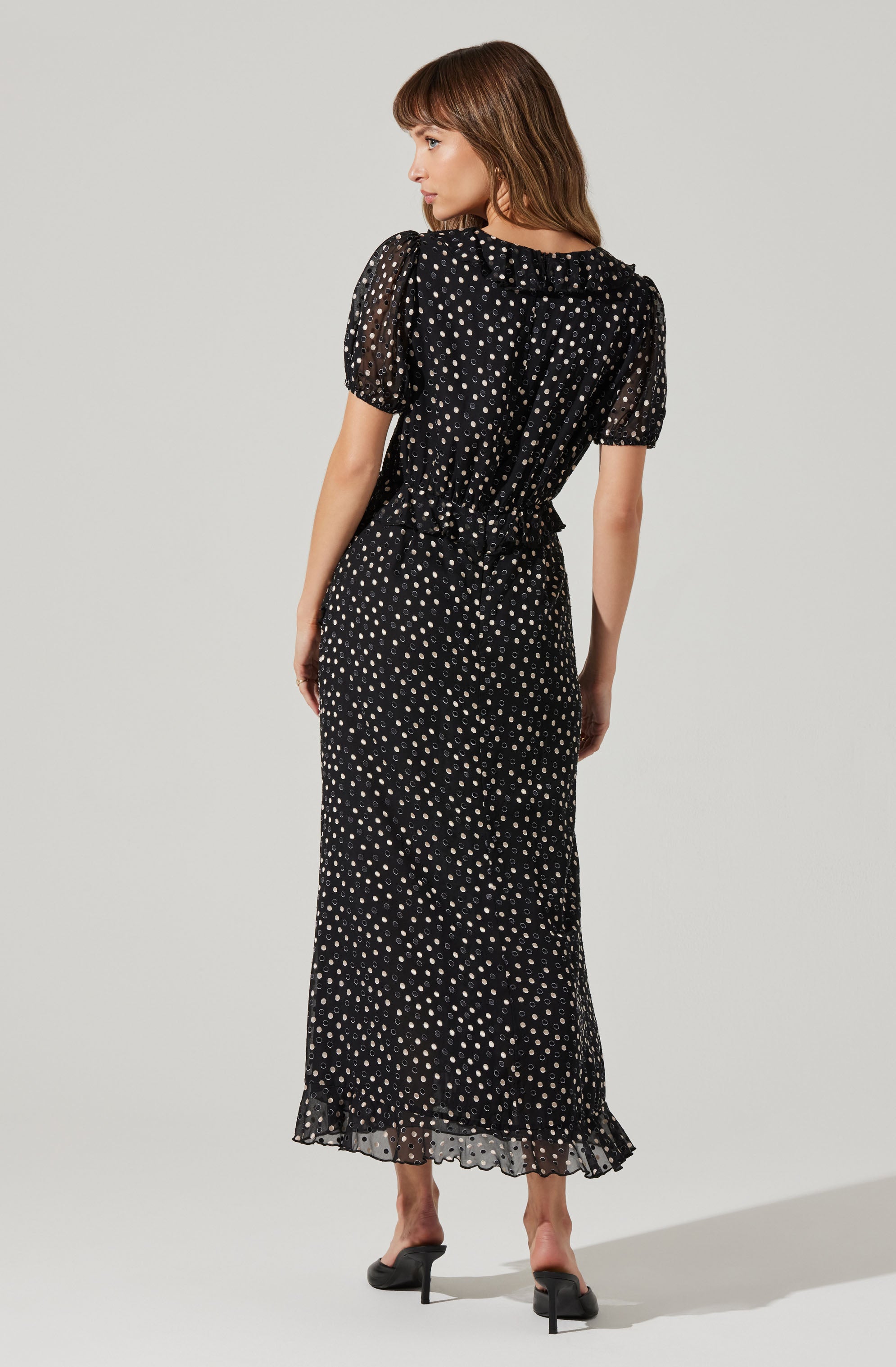 A woman is wearing a black, ankle-length dress with a white polka dot pattern. The dress has short, puffed sleeves and a ruffled collar at the neckline. The fabric appears lightweight and slightly sheer, with a smocked waistband that creates a gathered effect. She is also wearing black high-heeled shoes.