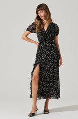 A woman is wearing a black, midi-length dress with a sheer, dotted pattern. The dress features a ruffled front with a tie at the neckline, short puffy sleeves, a high slit on the left side revealing her leg, and a ruffled hem. She is also wearing black high-heeled shoes.