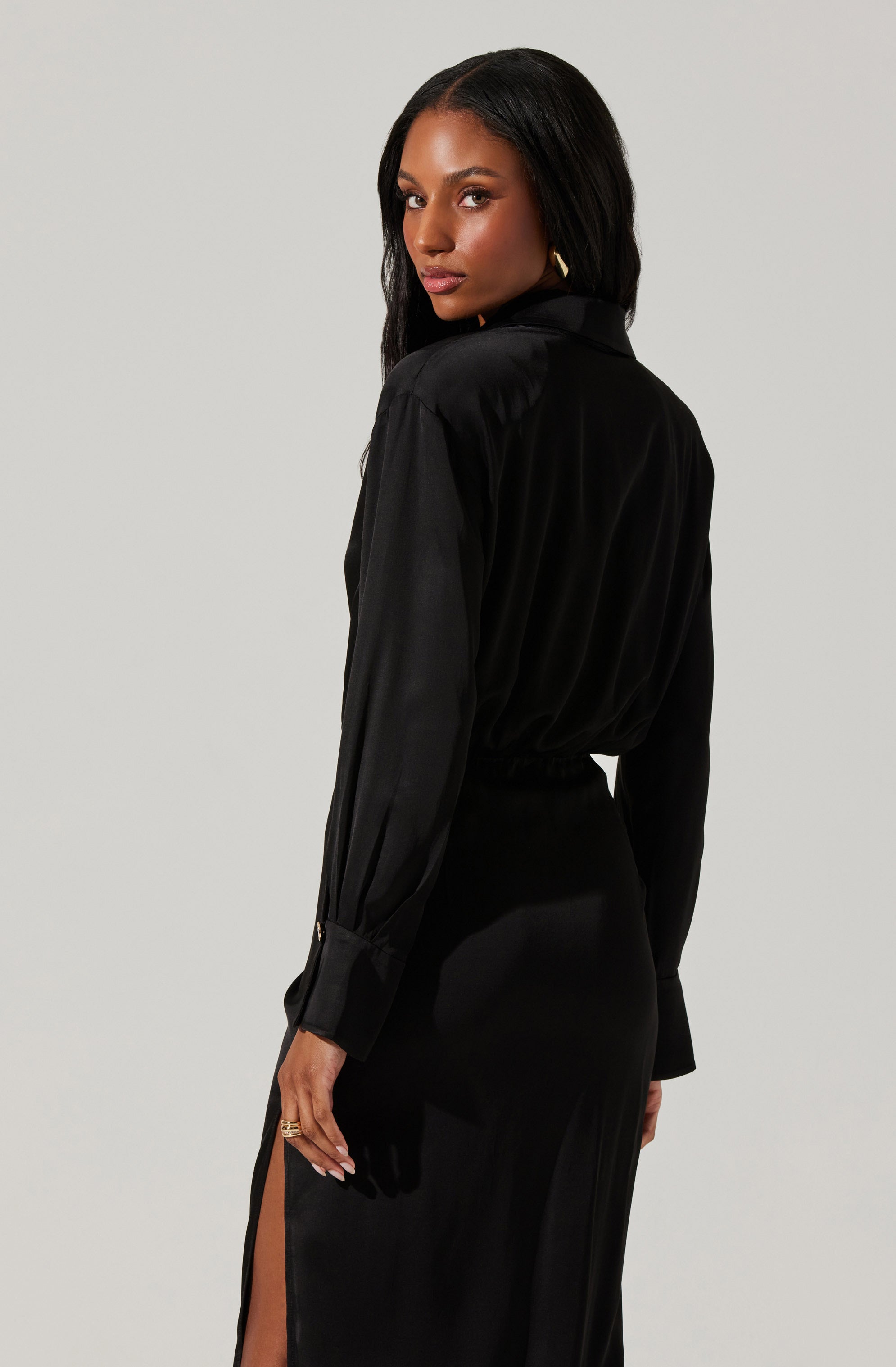 A woman is wearing a long, black satin dress with a slit on the side. The dress features a collared neckline, long sleeves with buttoned cuffs, and a fitted waist. The material has a shiny, smooth appearance.