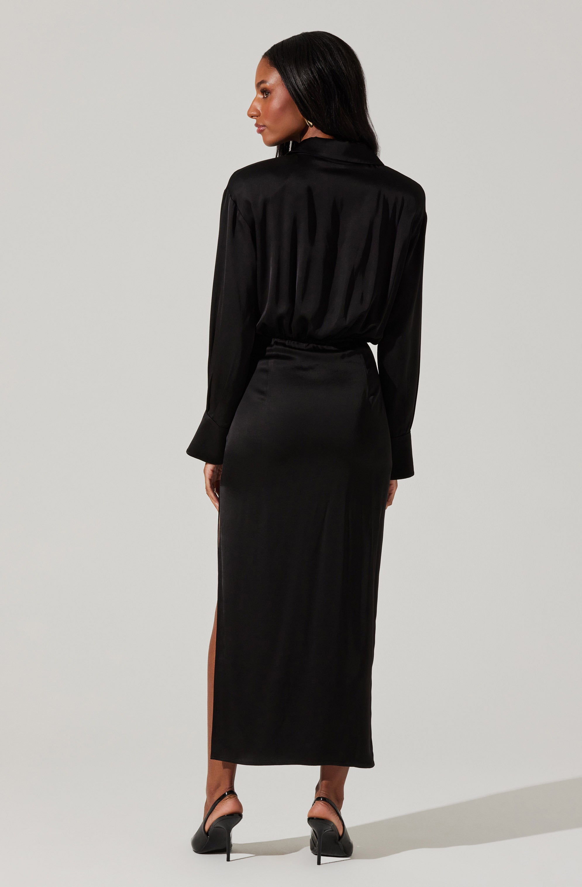 A woman is wearing a long, black satin dress with a high slit on the left side. The dress features long sleeves and a collar at the neckline. The fabric has a smooth, shiny texture and drapes elegantly. She is also wearing black high-heeled shoes.