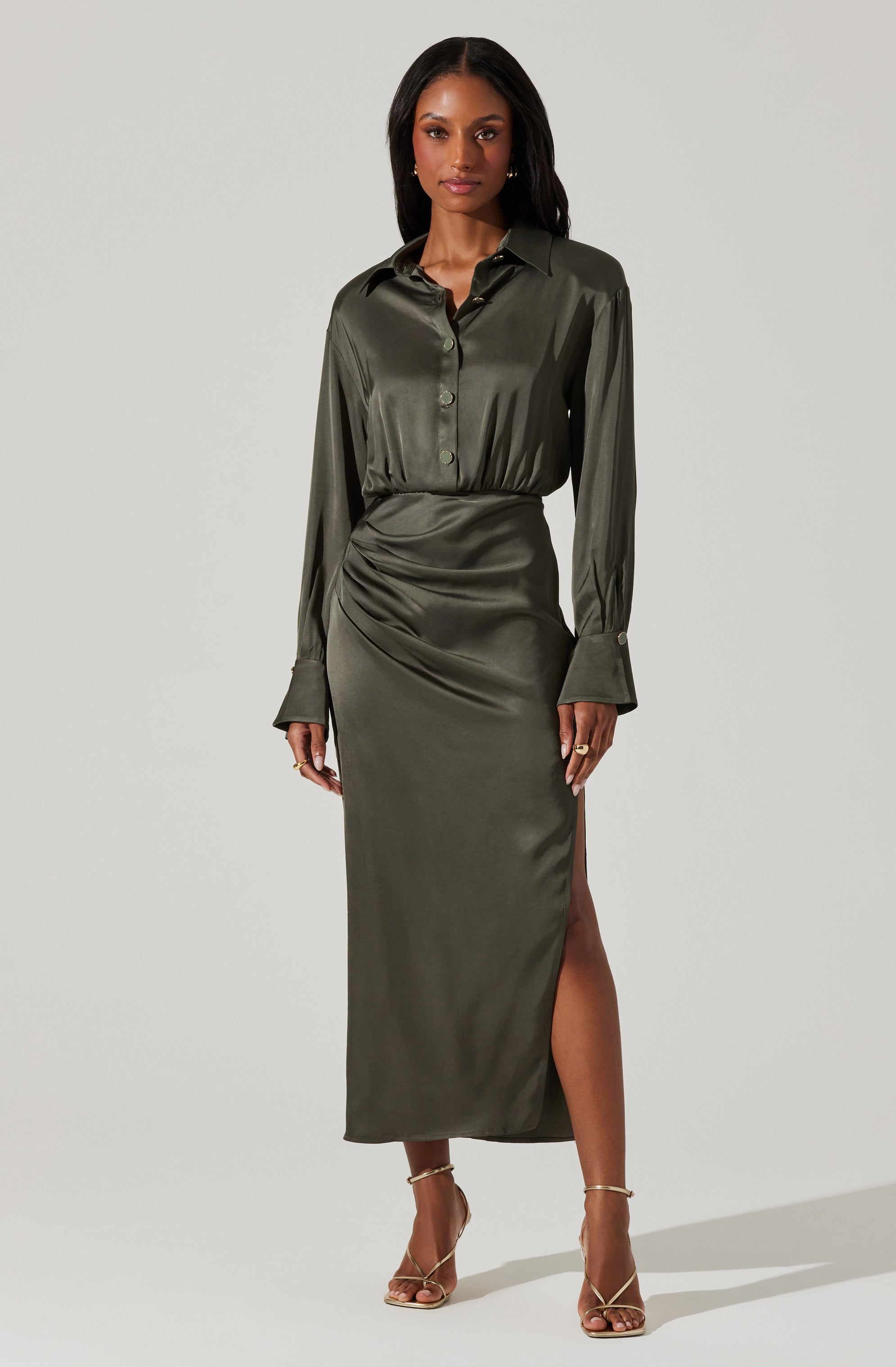 A woman is wearing a shiny, dark green satin outfit consisting of a long-sleeve button-up shirt with a collar and cuffs, tucked into a matching high-waisted, ankle-length skirt that features a side slit. She is also wearing strappy high-heeled sandals.