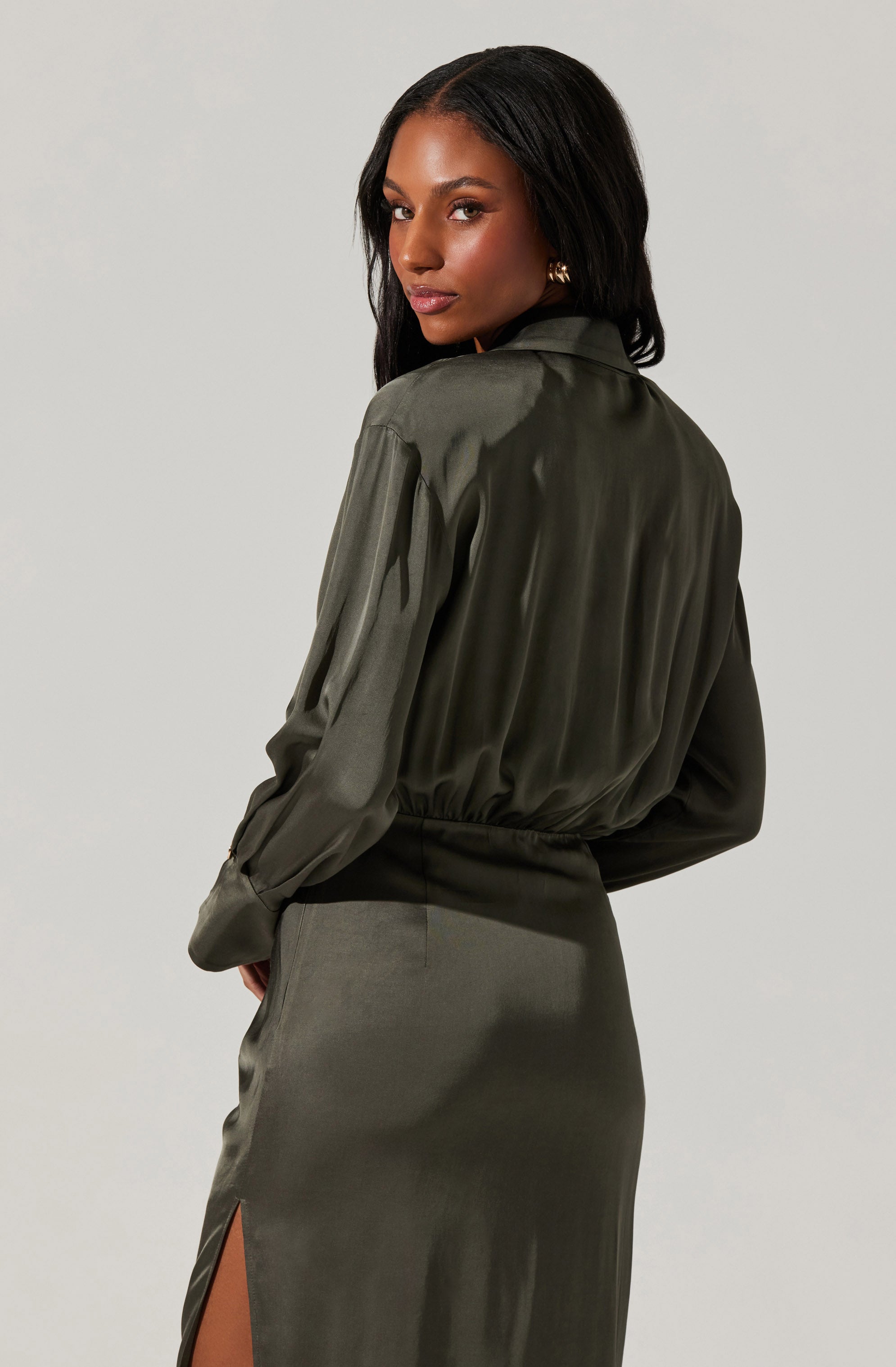 A woman is wearing an olive green satin blouse with long sleeves and a collar, tucked into a matching satin skirt that has a side slit.