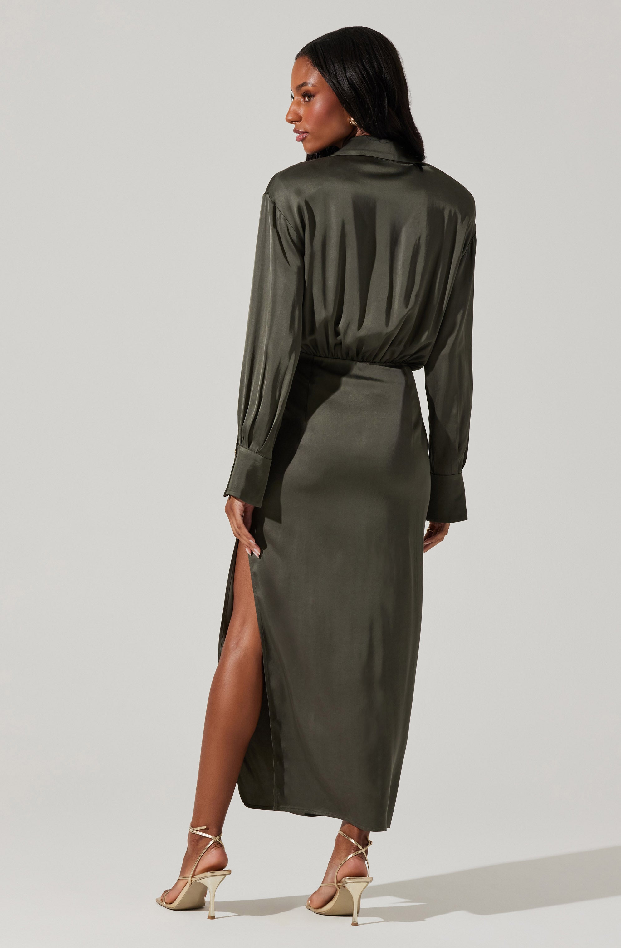 A woman is wearing a long, olive-green satin dress with a high slit on the side. The dress has long sleeves with wide cuffs and a belted waist. She is also wearing beige high-heeled sandals with ankle straps.