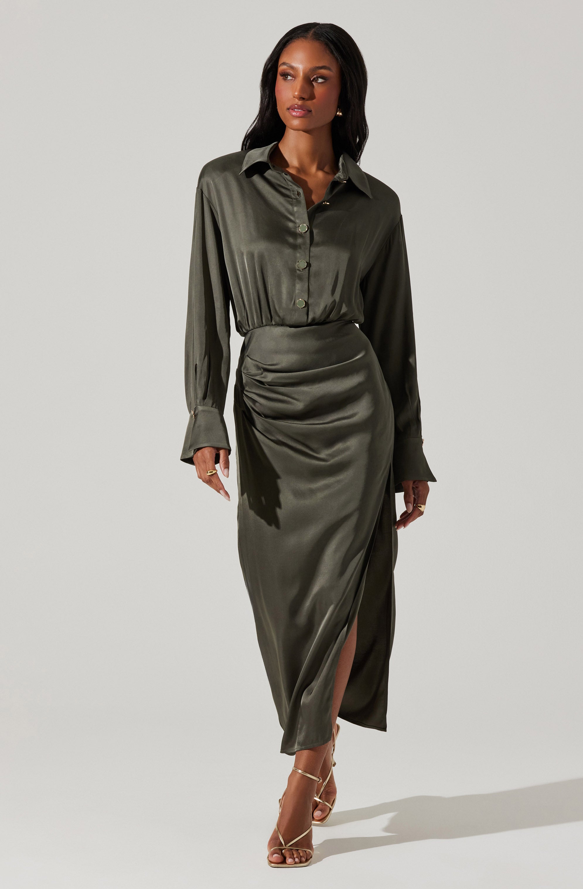 A woman is wearing an olive-green satin shirt with a collar and long sleeves, which are slightly loose and have cuffs. She pairs the shirt with a matching high-waisted satin skirt that features a front slit, draping details, and gathers on one side. She completes her outfit with strappy high-heeled sandals.