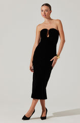 A woman is wearing a strapless, black velvet dress that is form-fitting and extends to mid-calf length. The dress features a keyhole cutout at the bust with gathered fabric around the bust area. She pairs the dress with black pointed-toe heels and gold accessories, including hoop earrings and a wide cuff bracelet.