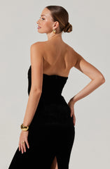 A woman is wearing a strapless black dress with a fitted silhouette and a slit on the side. She accessorizes with gold hoop earrings and a gold bracelet on her left wrist. Her hair is styled in a sleek, low bun.
