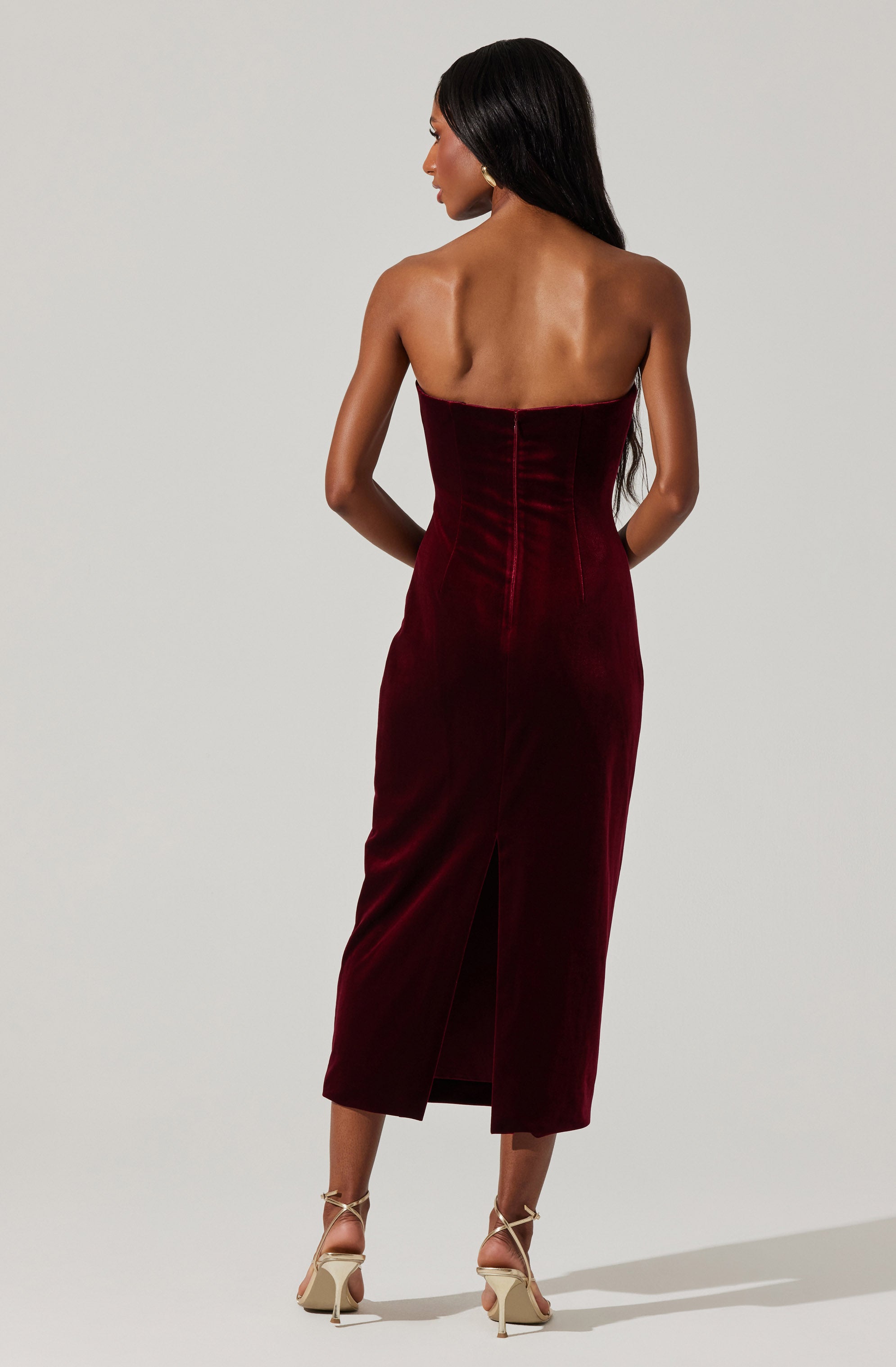 A woman is wearing a strapless, deep red velvet dress that is fitted at the waist and extends to mid-calf length. The dress features vertical seam details and a back slit at the hem. She pairs the outfit with beige strappy high-heeled sandals.