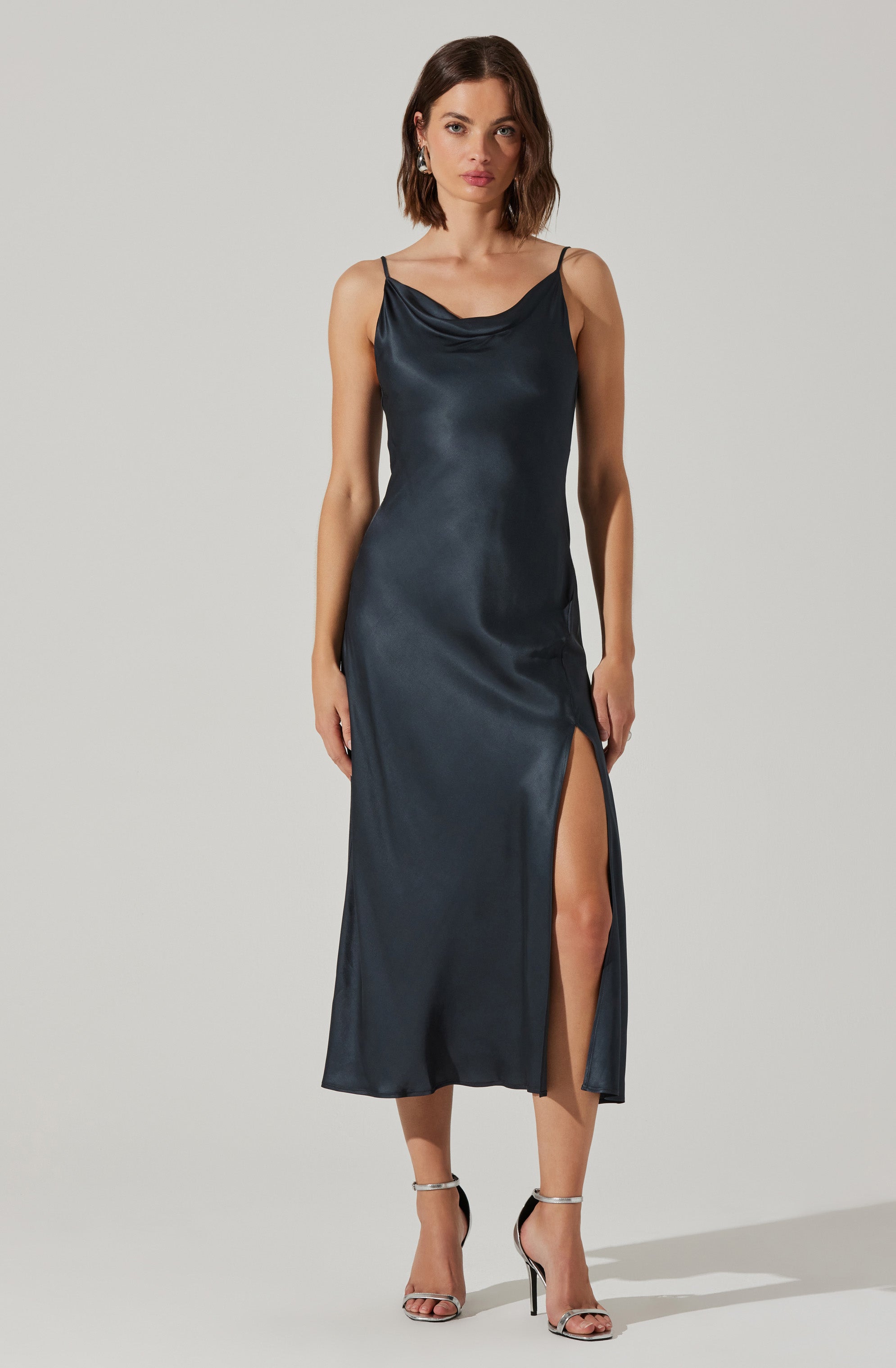A woman is wearing a dark satin slip dress with thin spaghetti straps. The dress features a cowl neckline and a high slit on the left side, revealing her leg. She is also wearing strappy high-heeled sandals.