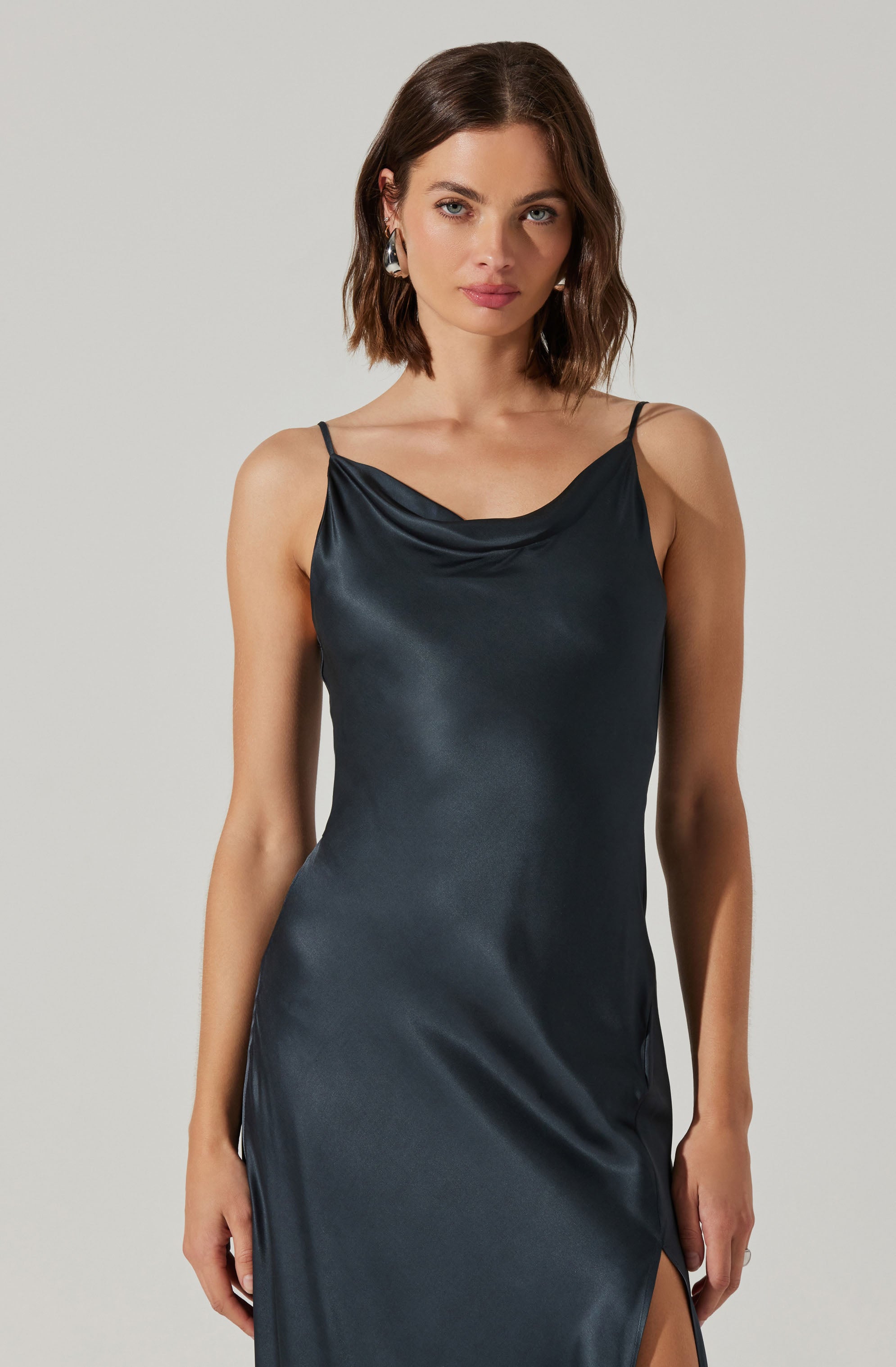 A woman is wearing a dark, silky slip dress with thin spaghetti straps. The dress features a cowl neckline and a side slit.
