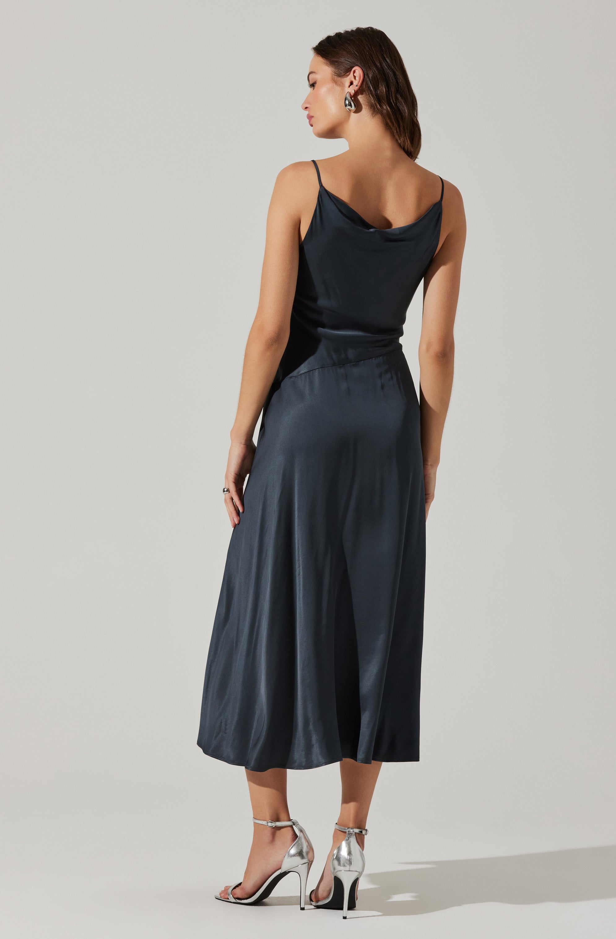 A woman is wearing a dark, silky, sleeveless dress with thin spaghetti straps. The dress has a cowl neckline at the back and a high-waisted, loose-fitting, midi-length skirt. She is also wearing shiny, metallic high-heeled sandals with ankle straps.