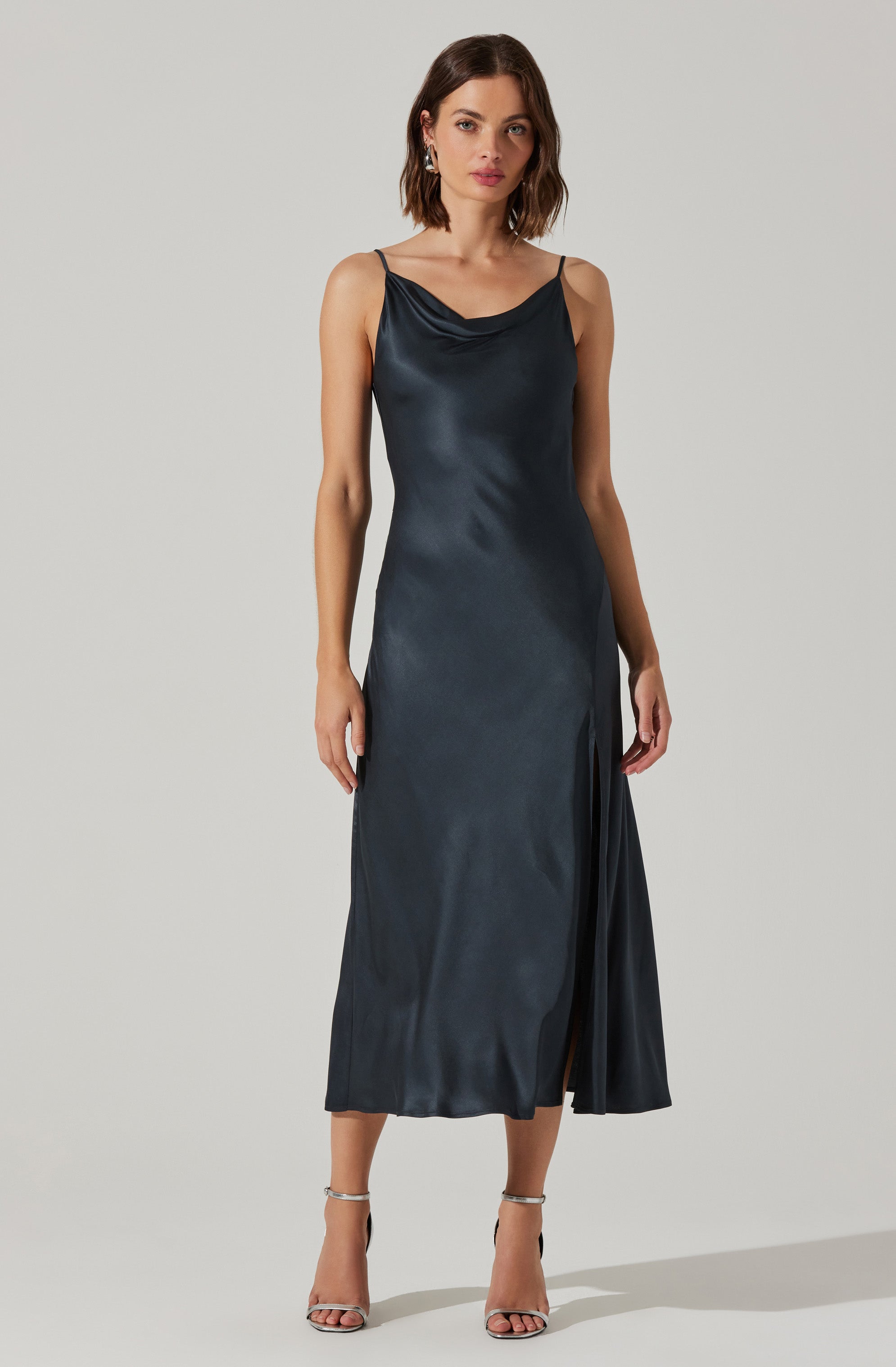 A woman is wearing a dark satin slip dress with thin spaghetti straps. The dress has a cowl neckline and a thigh-high slit on one side. She pairs the dress with silver high-heeled sandals.