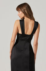 A woman is wearing a fitted black dress with wide shoulder straps that extend over the shoulders and down the back. The dress has a deep V-shaped back and subtle seam detailing. The fabric appears smooth and glossy.