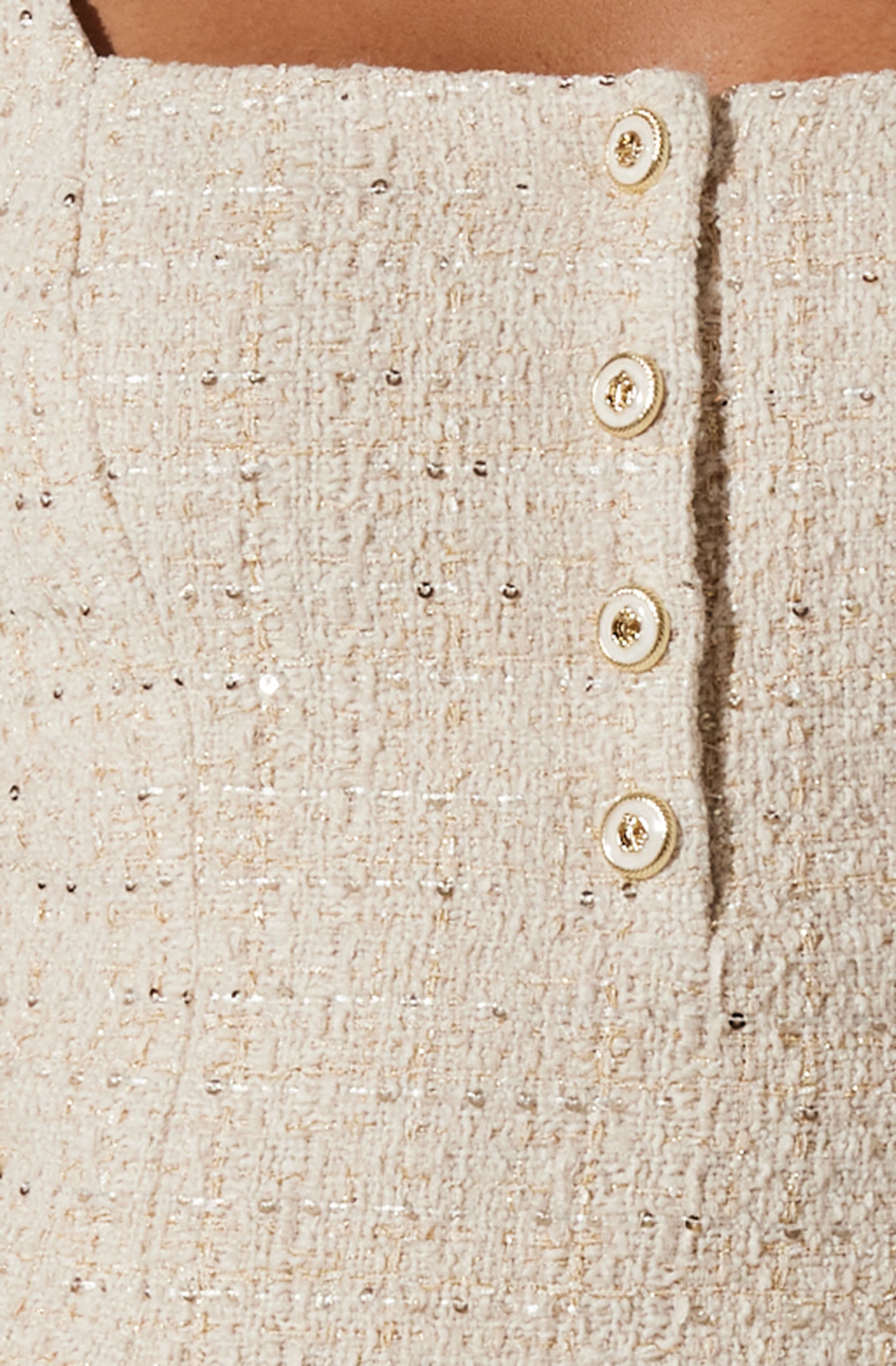 A close-up of a light-colored, textured, tweed fabric skirt with four decorative buttons aligned vertically on the front. The buttons are round, with a pearl-like appearance, and feature a small, embedded gemstone at the center. The skirt has a subtle shimmer with small metallic threads woven throughout.