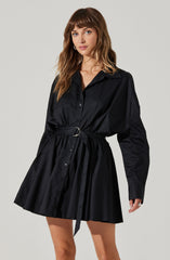 A woman is wearing a black, long-sleeved shirt dress made from a shiny, satin-like fabric. The dress features a pointed collar, button-down front, and a matching belt with a circular metal buckle at the waist. The skirt portion of the dress is gathered and flares out slightly, ending above the knees.