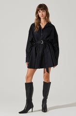 A woman is wearing a black, long-sleeved shirt dress with a collar and buttons down the front. The dress features a gathered waist with a matching fabric belt that has a silver buckle. She is also wearing tall black leather boots that reach just below the knees and have pointed toes and high heels.