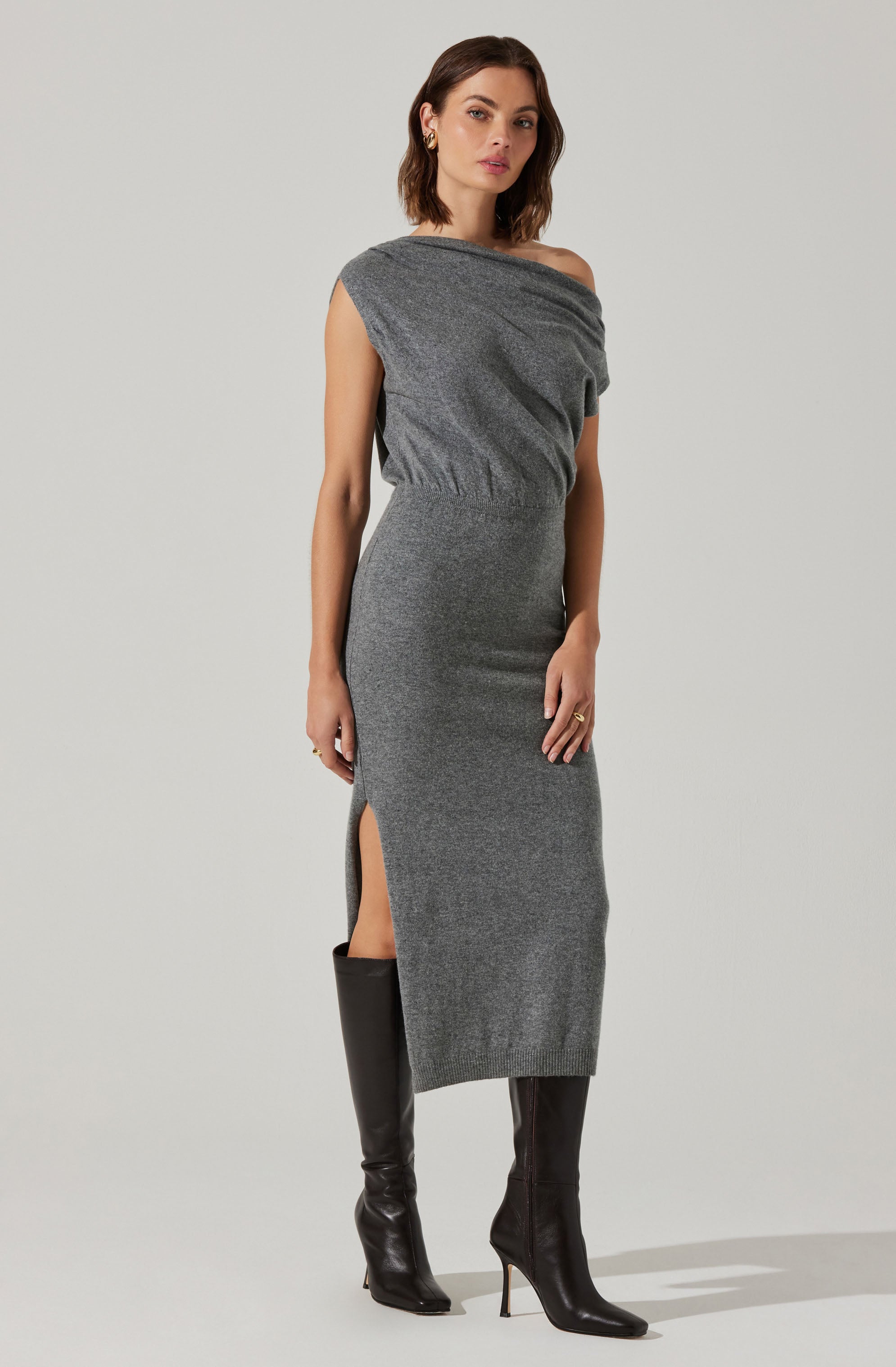 A woman is wearing a gray, one-shoulder, midi-length dress with a side slit. The dress appears to be made of a soft, knit fabric that fits snugly around her body. She pairs the dress with tall black heeled boots.