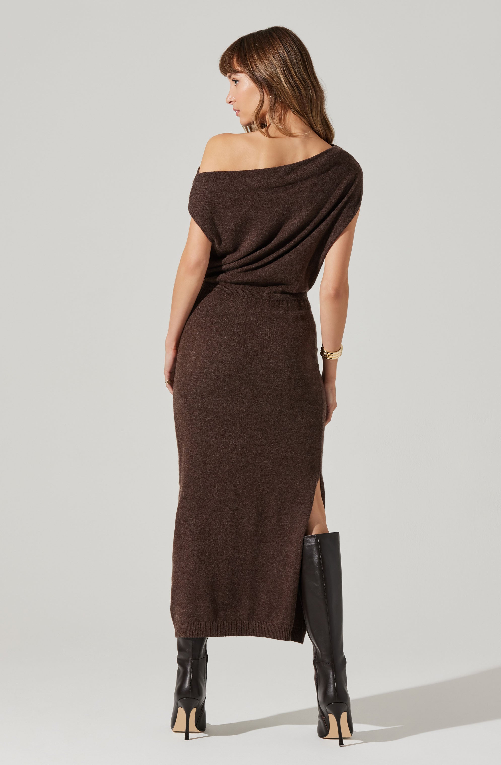 The woman is wearing an off-the-shoulder, short-sleeve top in a dark brown knit fabric, paired with a matching high-waisted, ankle-length skirt with a side slit. She is also wearing black high-heeled boots and a gold bracelet on her left wrist.